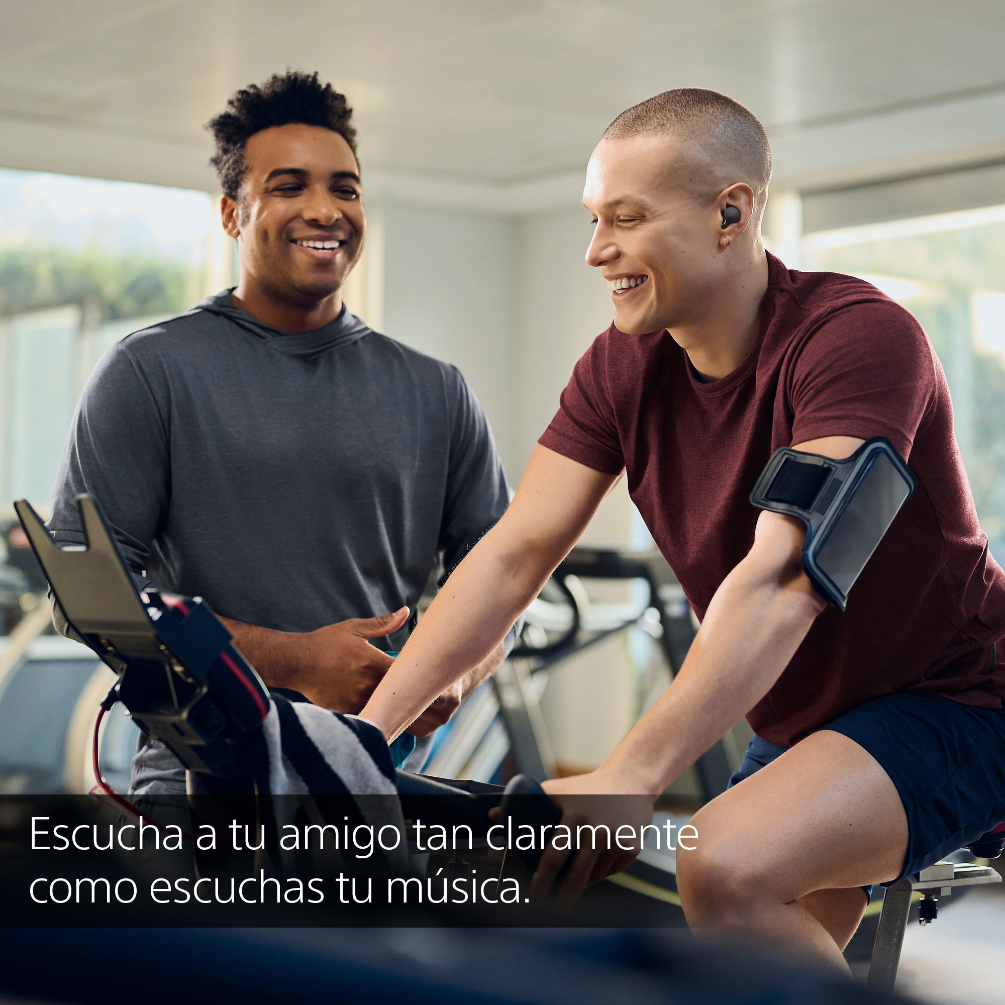 Dos hombres en el gimnasio sonriendo, uno en bicicleta estática, con texto en español.