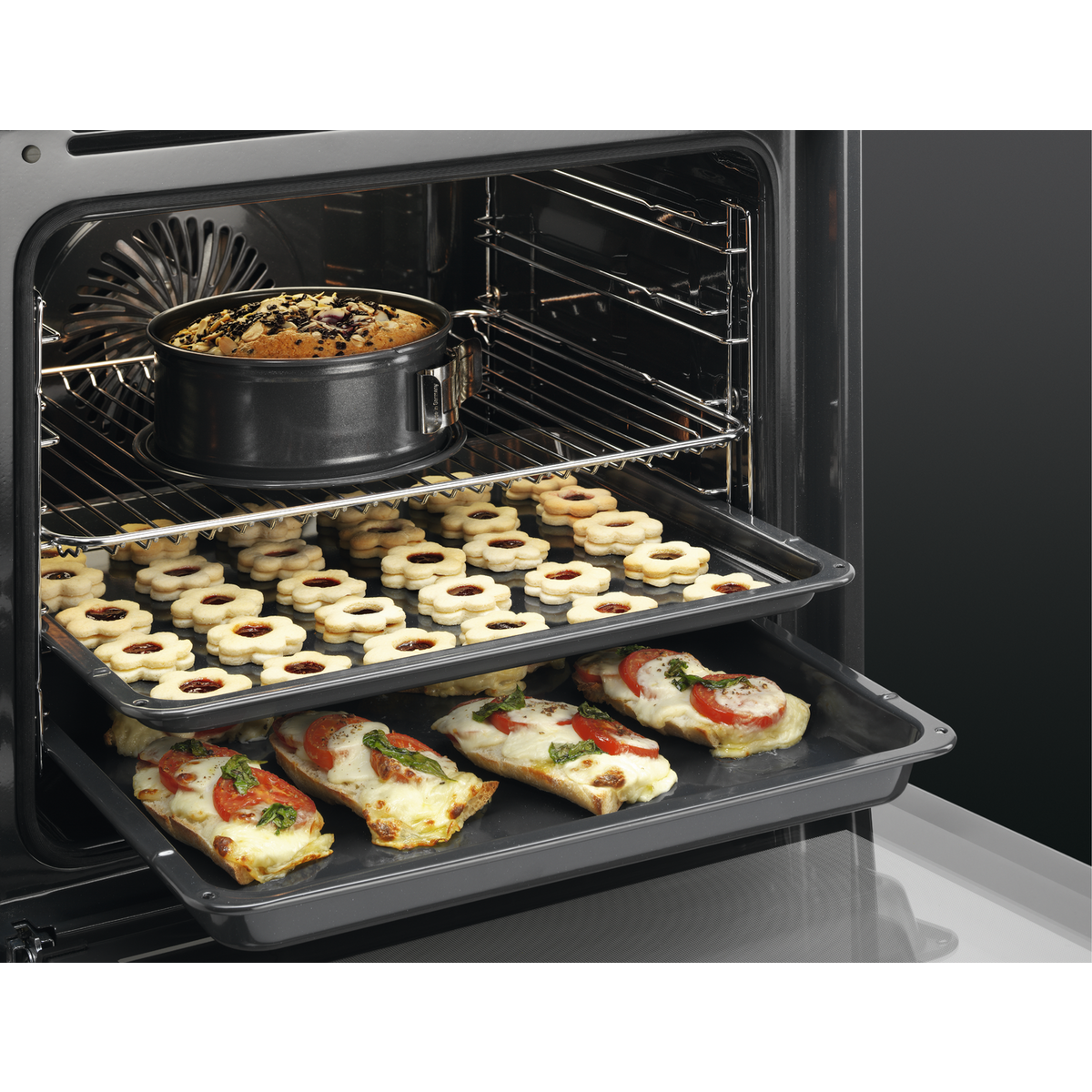 Een open oven met drie trays met eten. Boven: cake in een pan. Midden: koekjes. Onder: pizza-achtig brood.