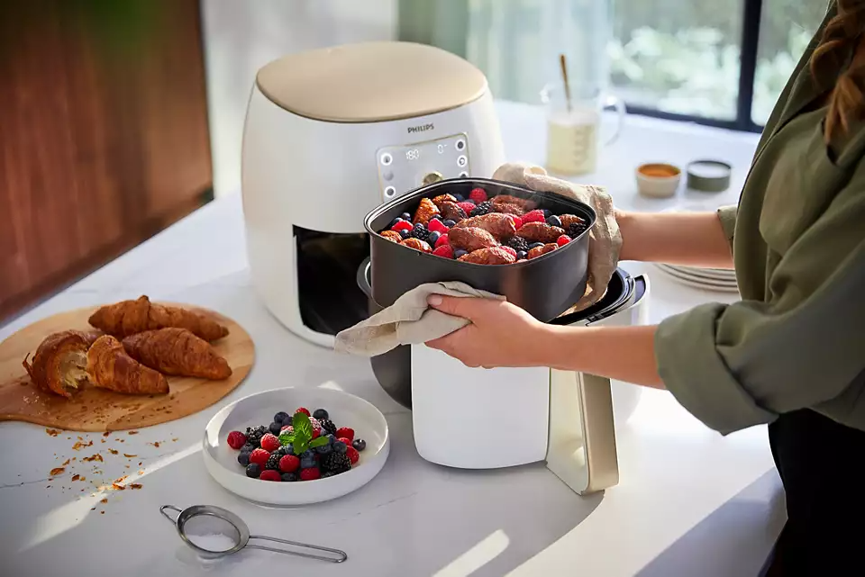 Egy személy egy forrólevegős sütőt tart főtt étellel és bogyókkal. A asztalon croissant, egy bogyós tál és fűszerek is vannak.