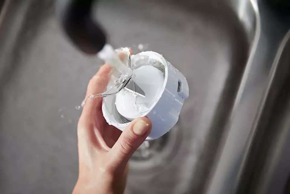 Egy kéz öblíti a fehér konyhai robotgép tartozékát folyó víz alatt a mosogatóban.