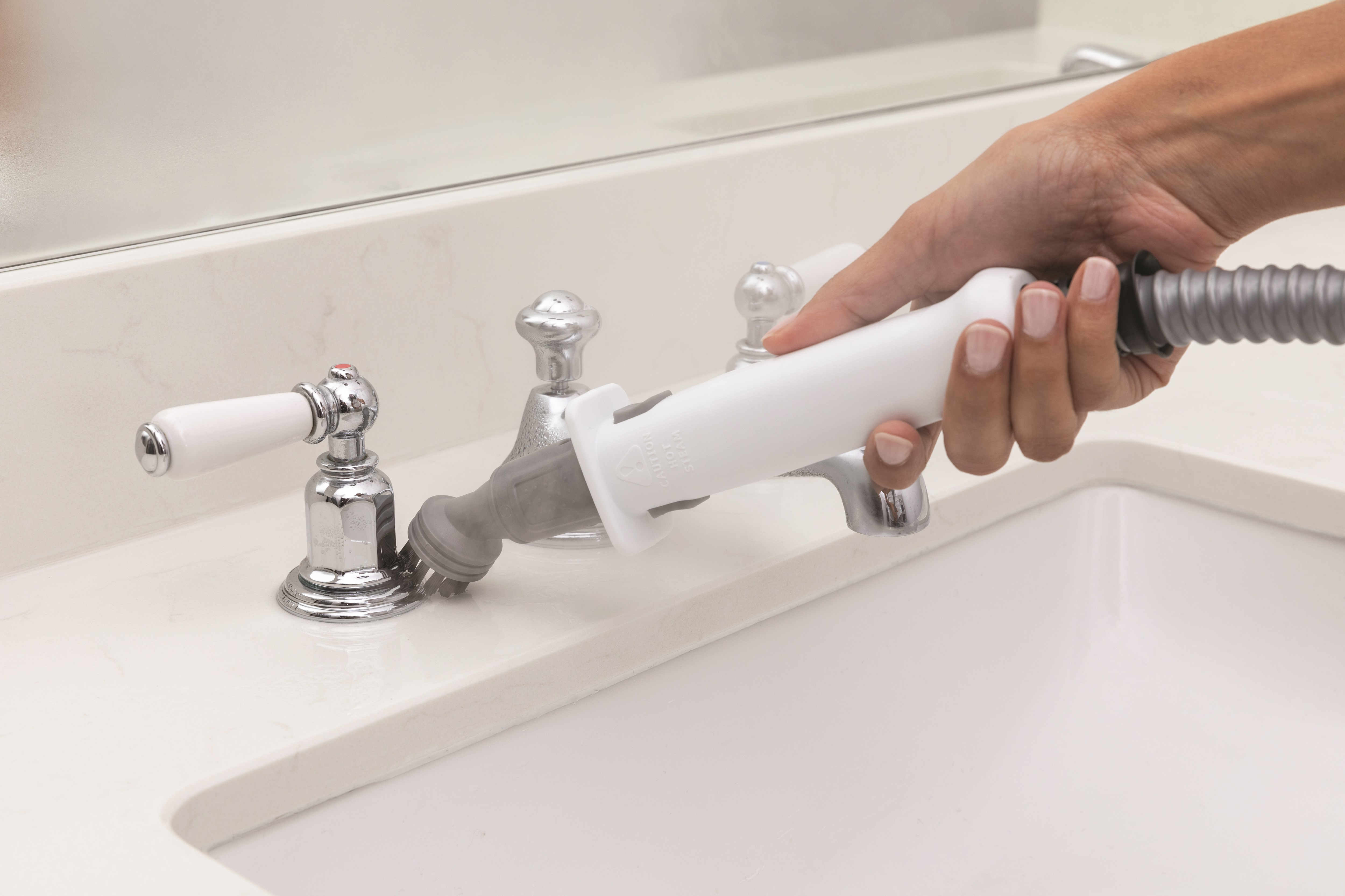 Een hand maakt een kraan schoon met een schoonmaaktool. De wastafel en kraan zijn wit.