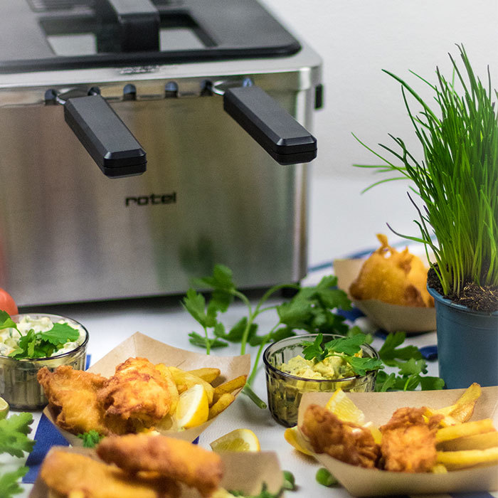 Piatti di pesce fritto e patatine fritte con fettine di limone, guacamole e erba cipollina, vicino a una friggitrice.