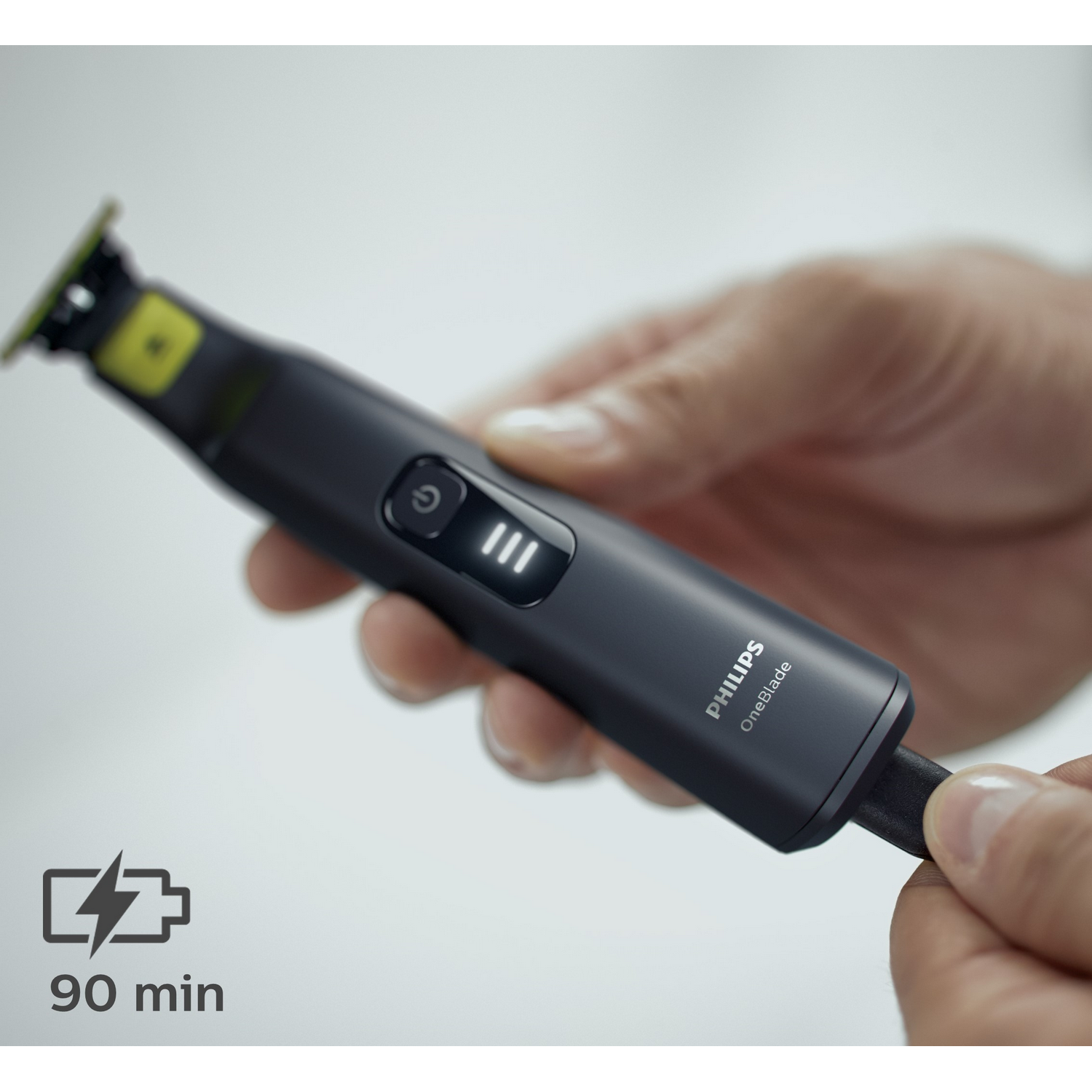 Una persona sostiene un recortador Philips OneBlade negro y gris. La imagen incluye un indicador de batería de 90 minutos.