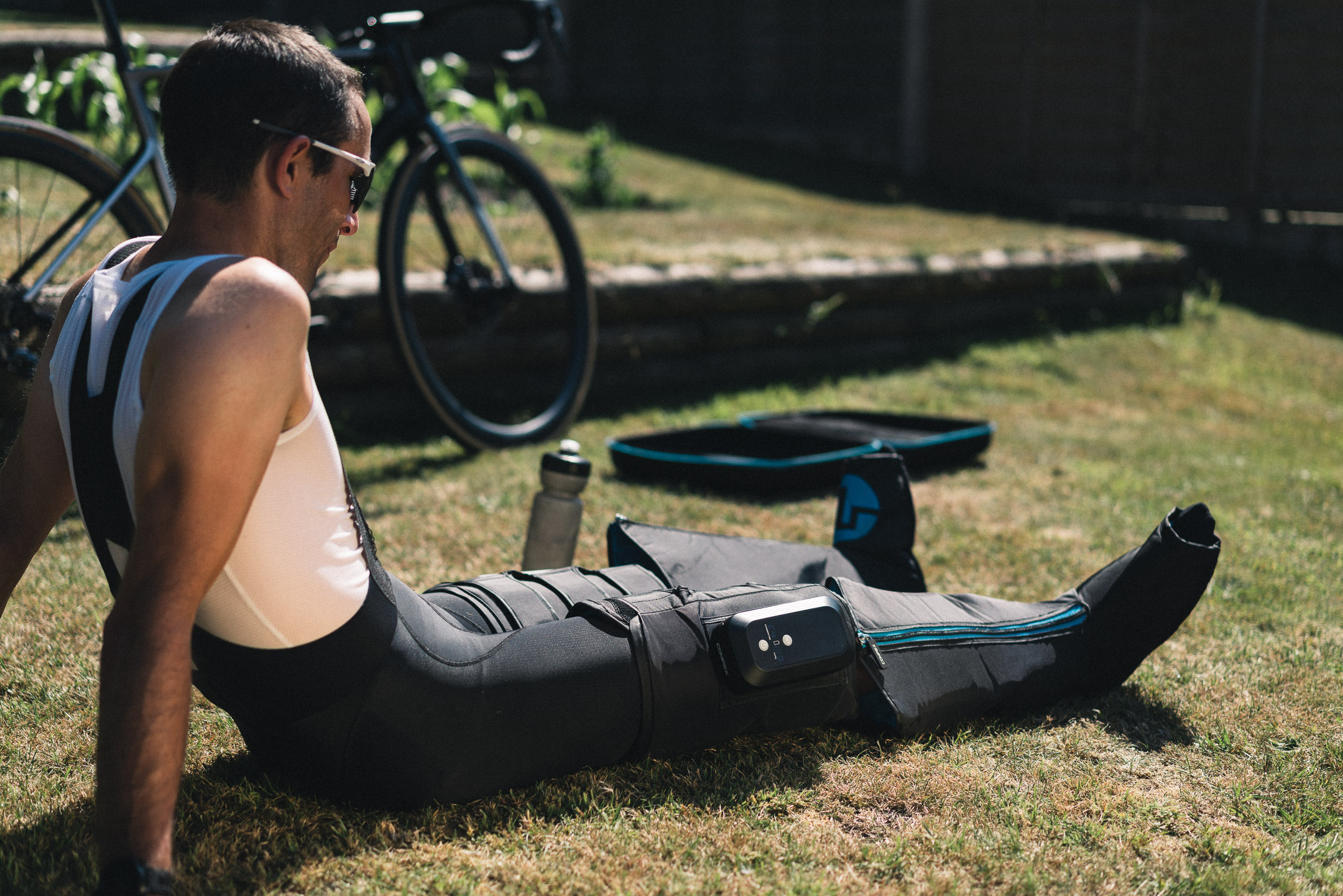 Ein Mann in Sportkleidung mit Bein-Erholungsgeräten auf Gras, Fahrrad im Hintergrund.