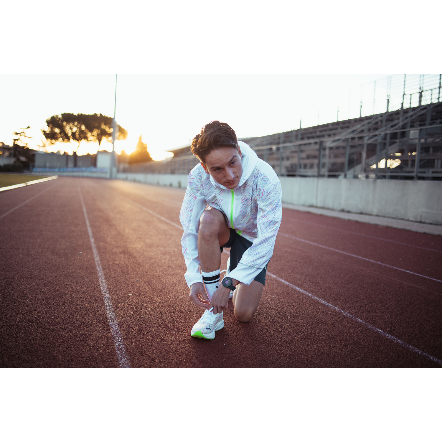 Atleta atándose los zapatos de correr en una pista, día soleado, chaqueta y pantalones cortos blancos.
