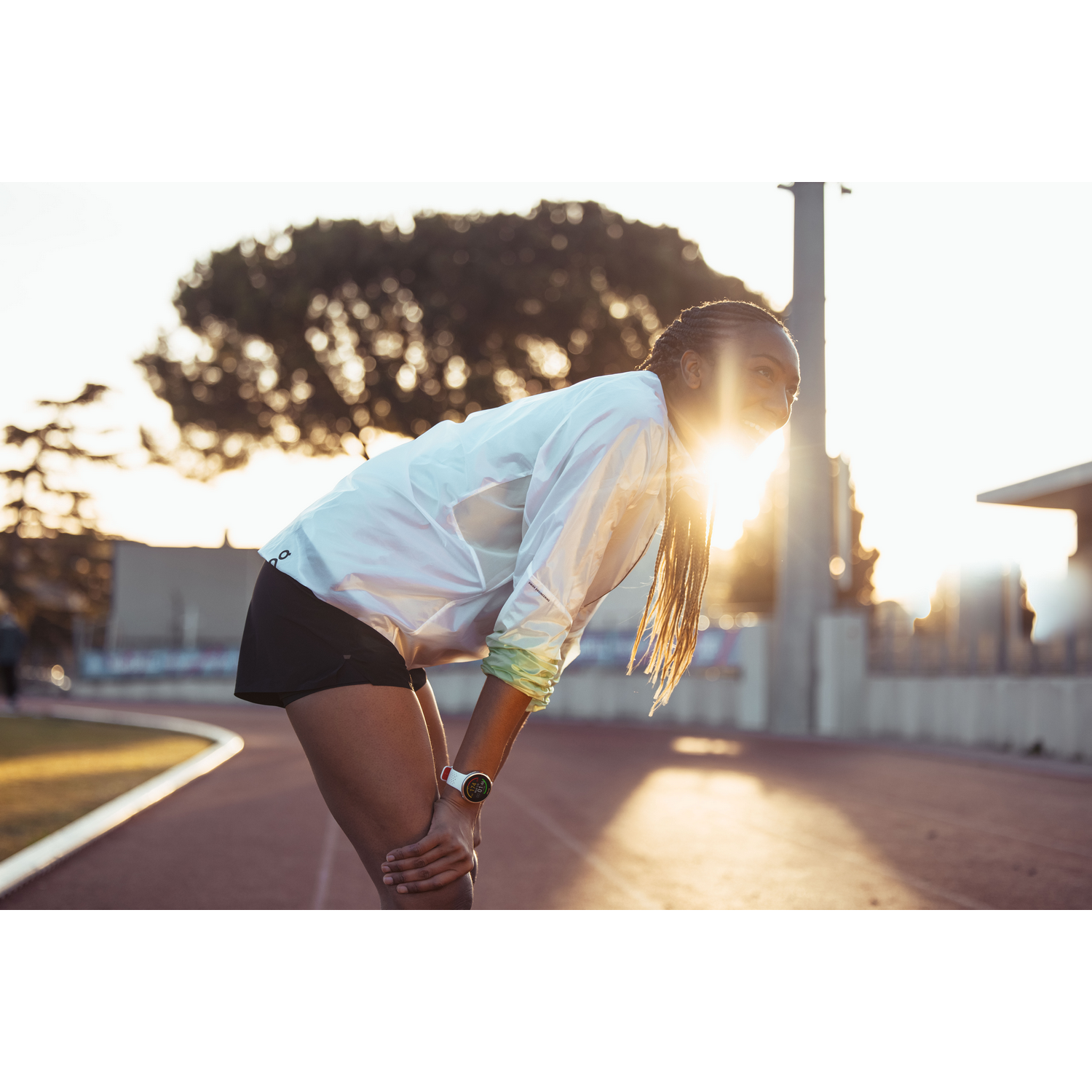 Una mujer en ropa deportiva se agacha, con las manos en las rodillas, en una pista; fondo soleado.