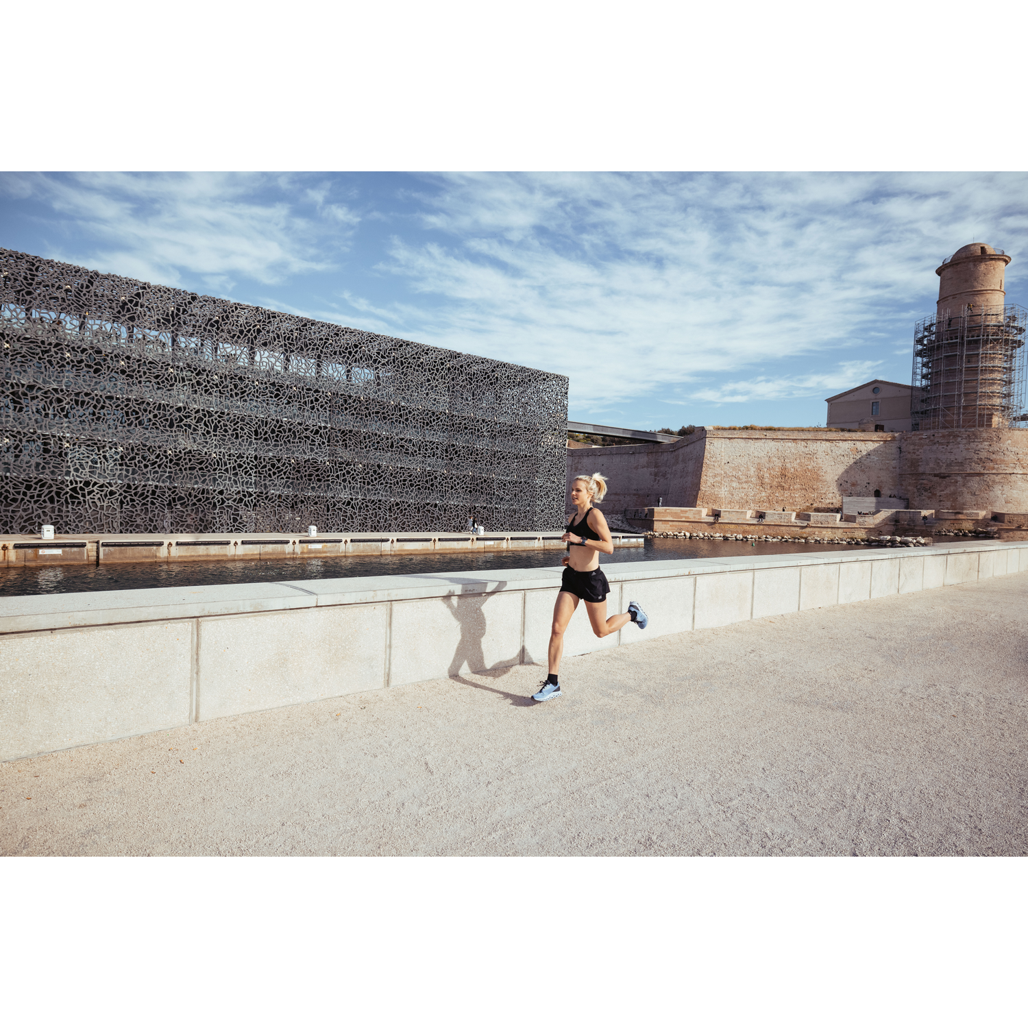 Mujer rubia corriendo cerca de un edificio moderno y un muro de piedra en un día soleado.