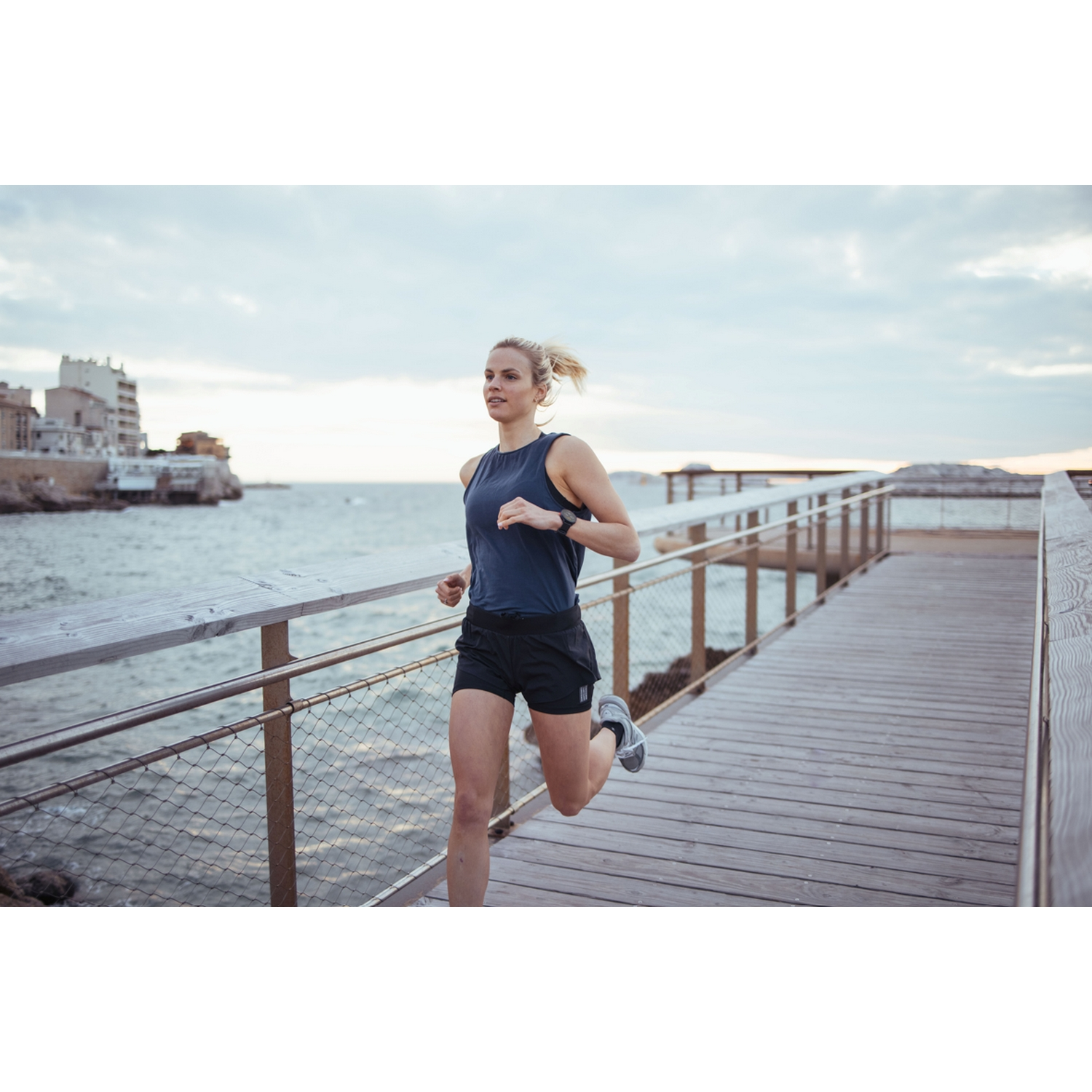 Mujer corriendo en un muelle de madera junto al mar, usando ropa deportiva, en un entorno costero escénico.