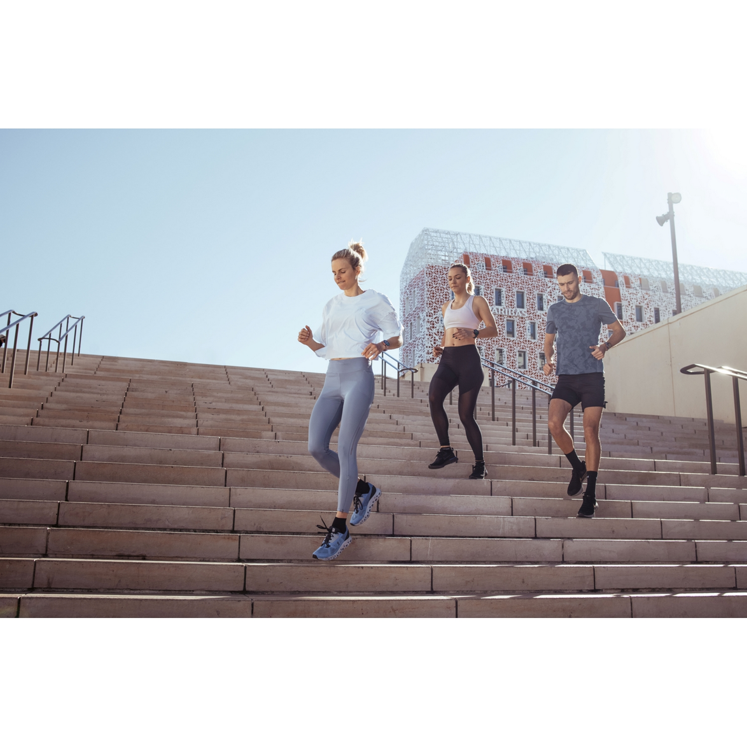 Tres personas corriendo por las escaleras. Una mujer delante, dos personas detrás. Cielo azul despejado al fondo.