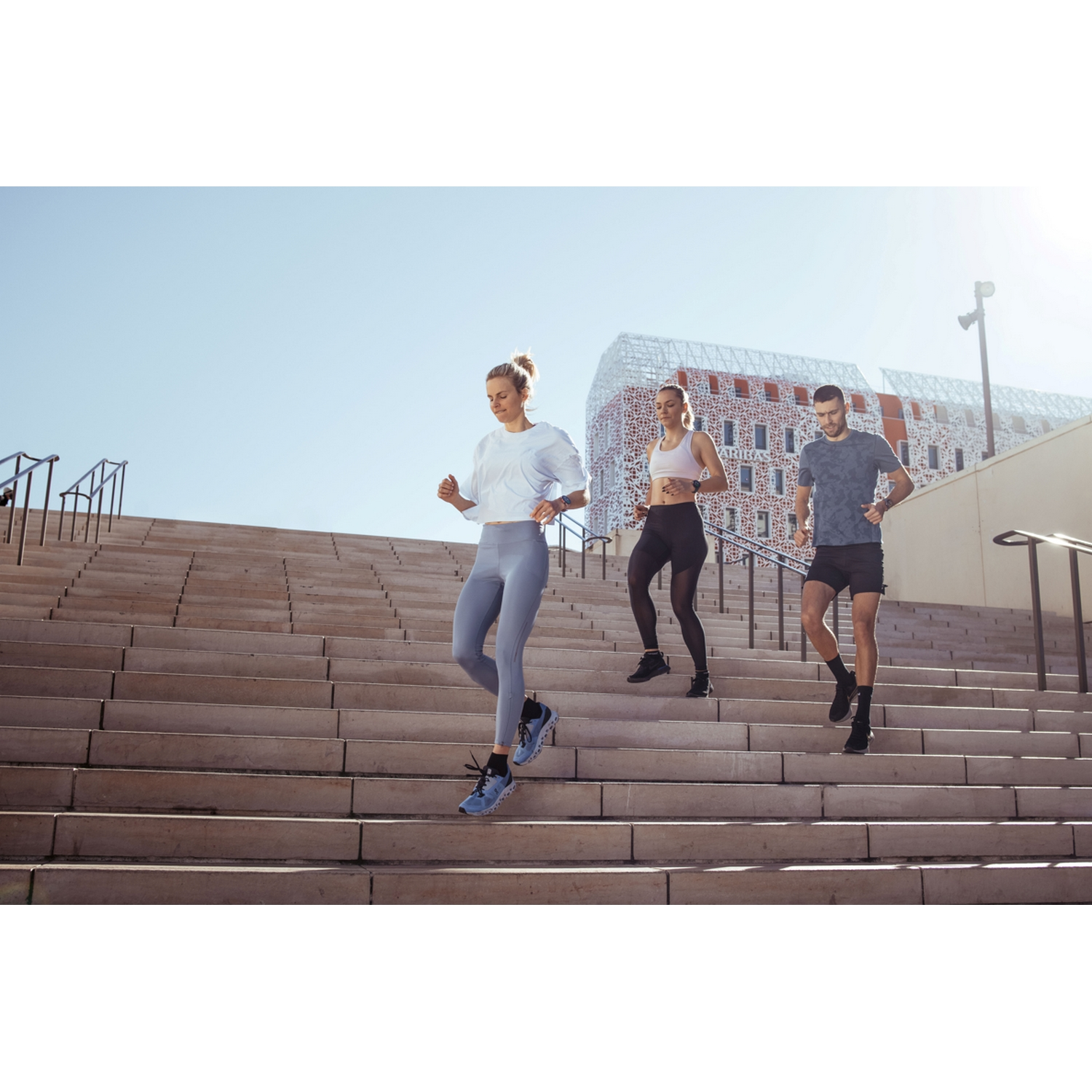 Tres personas corriendo por escaleras al aire libre. El cielo es azul y el sol brilla.