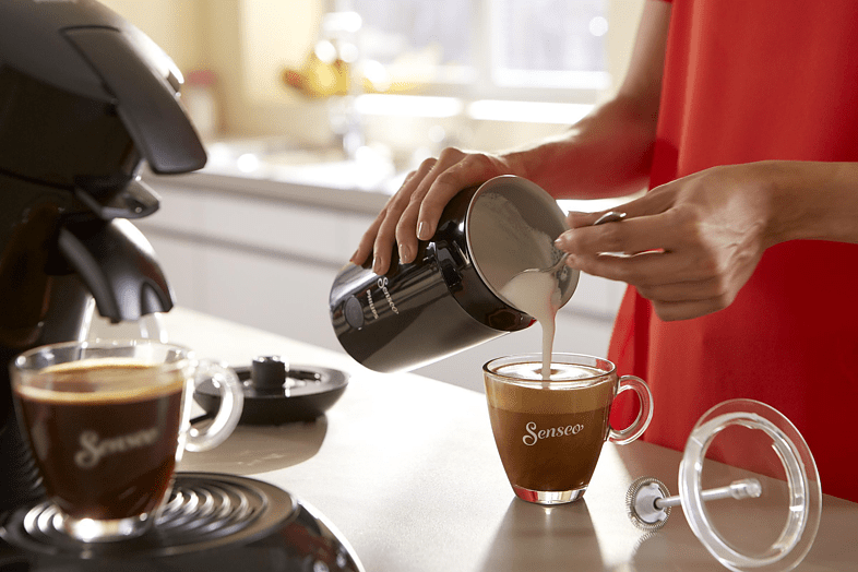 Een persoon giet melk in een glas koffie, andere koffie en een opschuimer zijn zichtbaar.