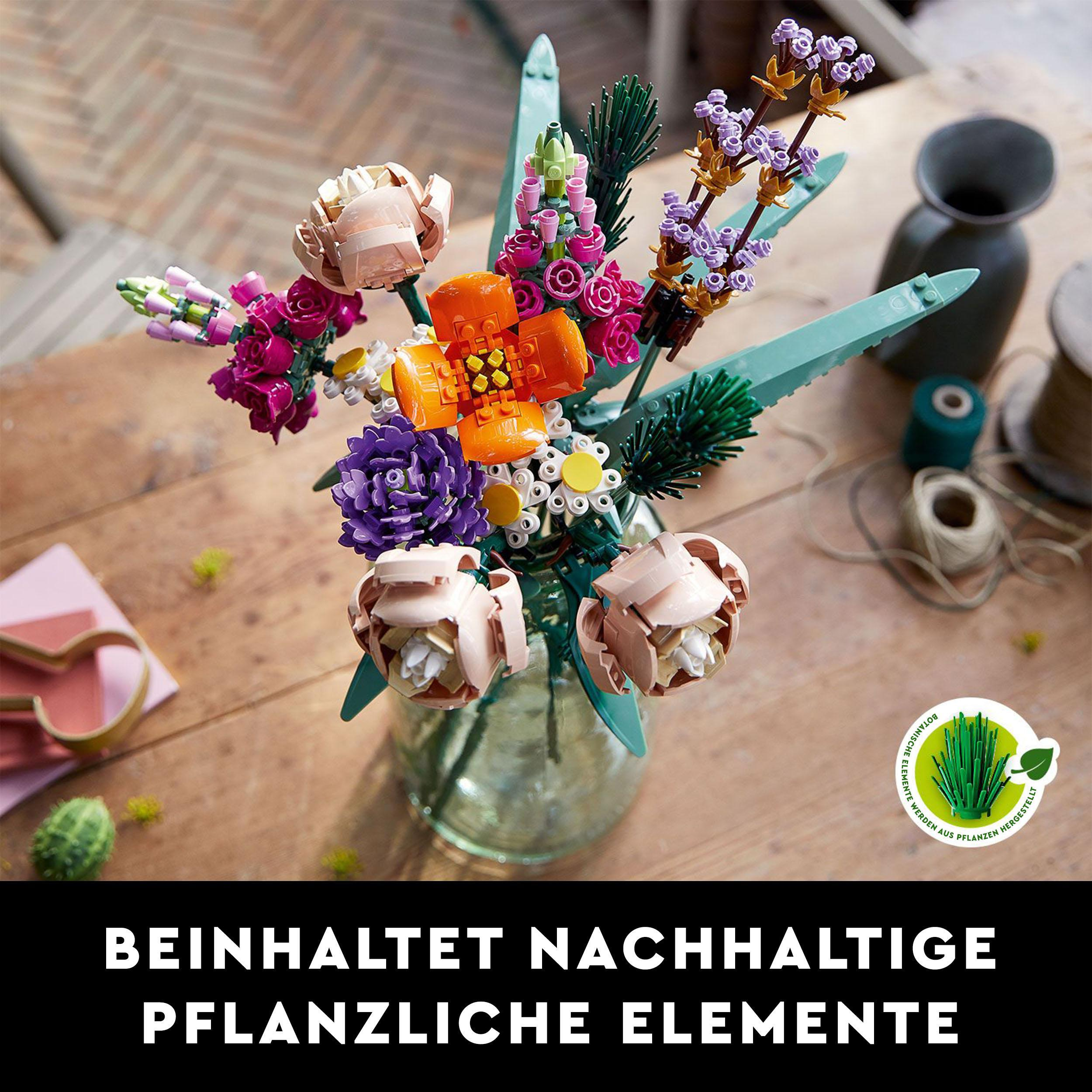 Ein Strauß bunter Lego-Blumen in einer Vase auf einem Holztisch. Grüne Blätter und rosa Blütenblätter.