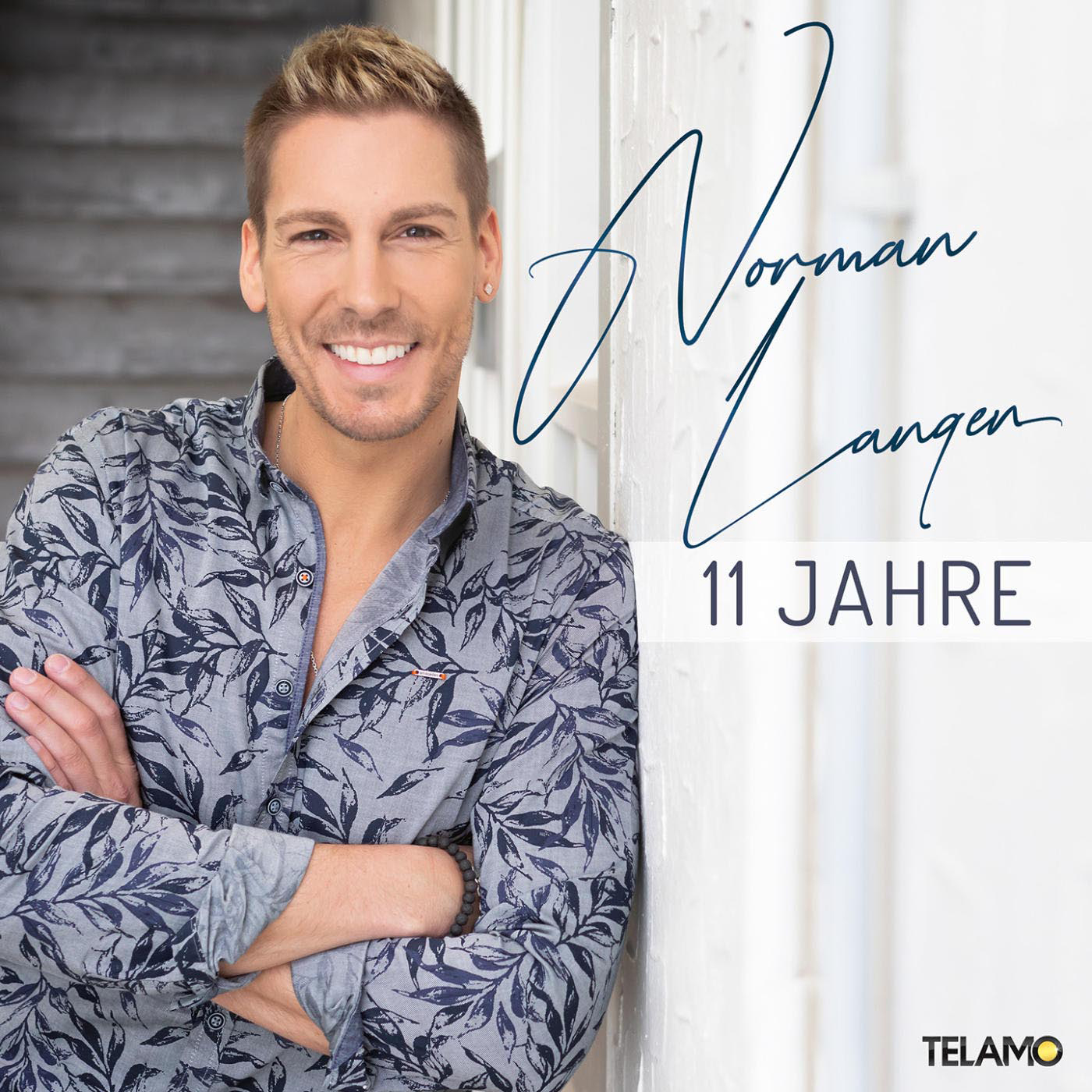 Ein Mann in einem gemusterten Hemd lehnt an einer Wand und lächelt.