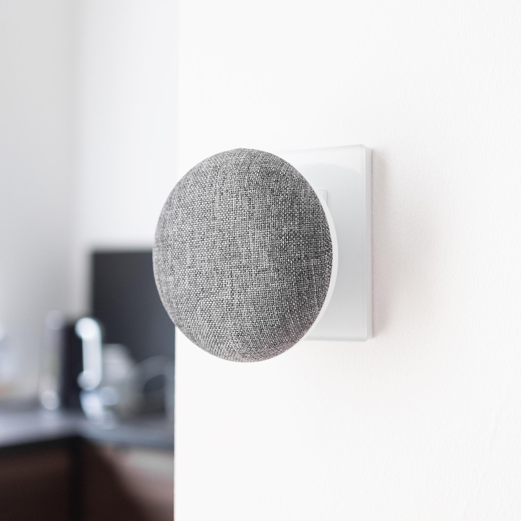 Ein grauer Google Nest Mini-Lautsprecher, montiert an einer weißen Wand mit Halterung.