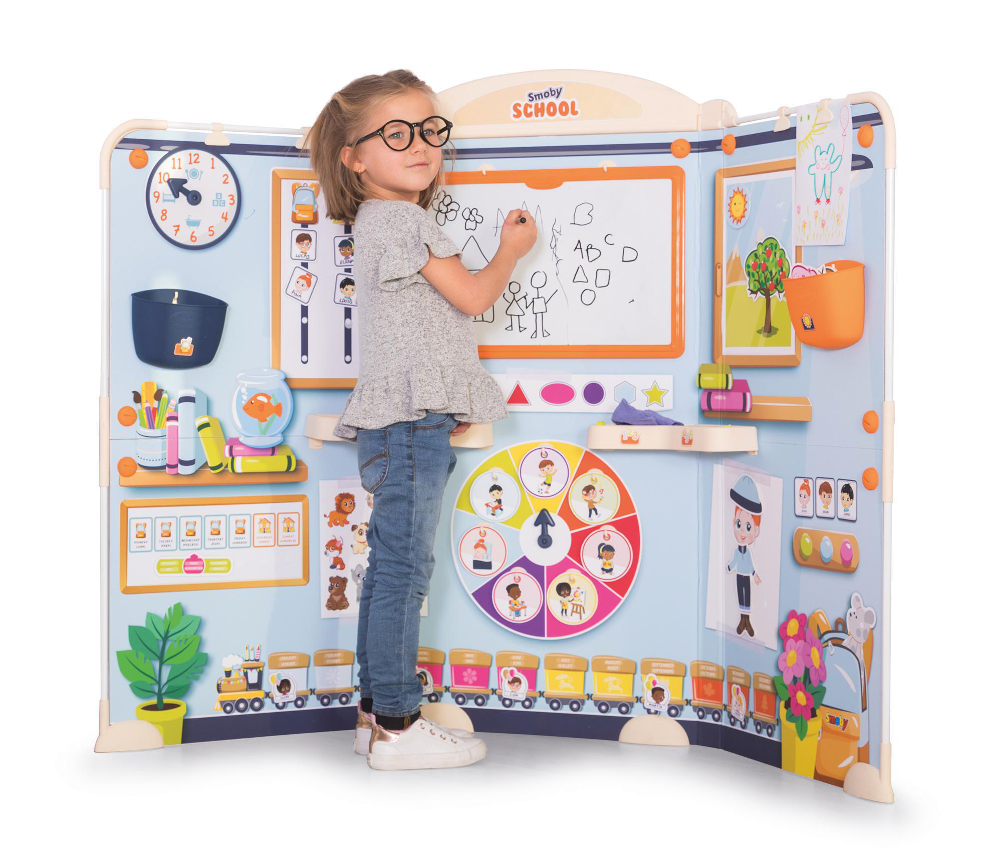 Junges Mädchen mit Brille zeichnet auf einer Schultafel mit Uhr, Fischglas und Lernwerkzeugen.