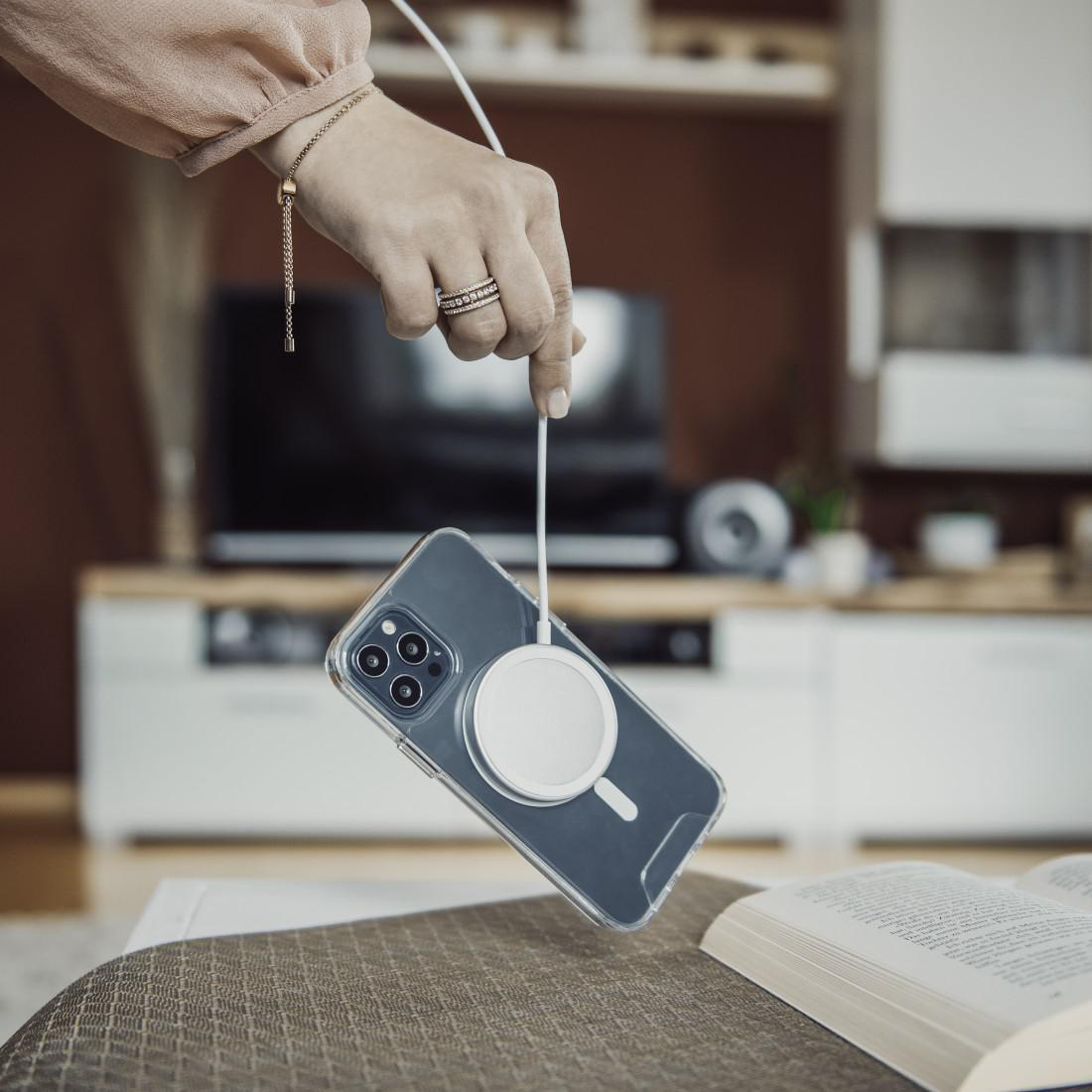 Eine Hand, die ein Ladekabel hält, das mit einem Telefon verbunden ist. Tisch und Fernseher im Hintergrund.