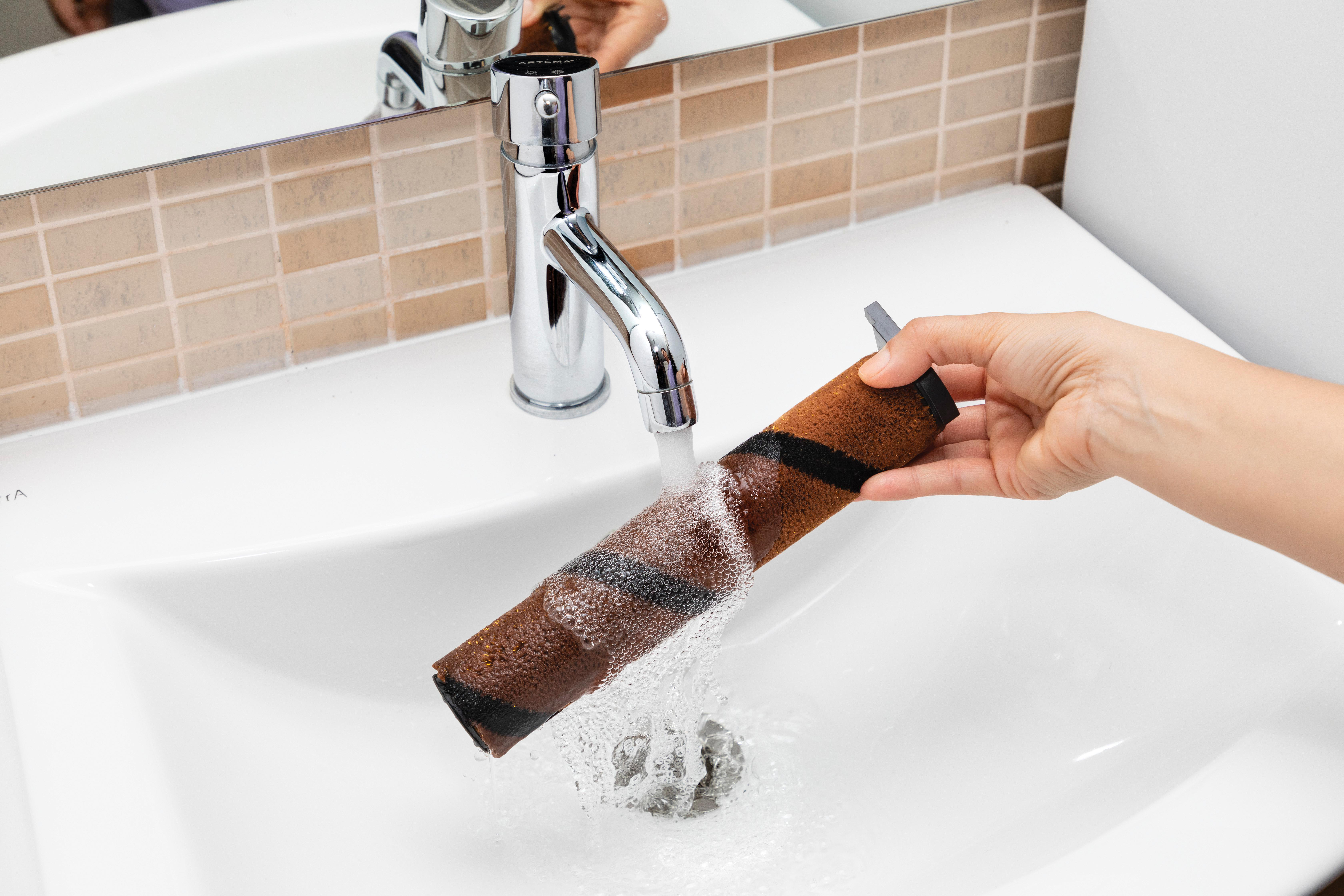 Eine Person wäscht einen Filter unter einem laufenden Wasserhahn in einem weißen Waschbecken.