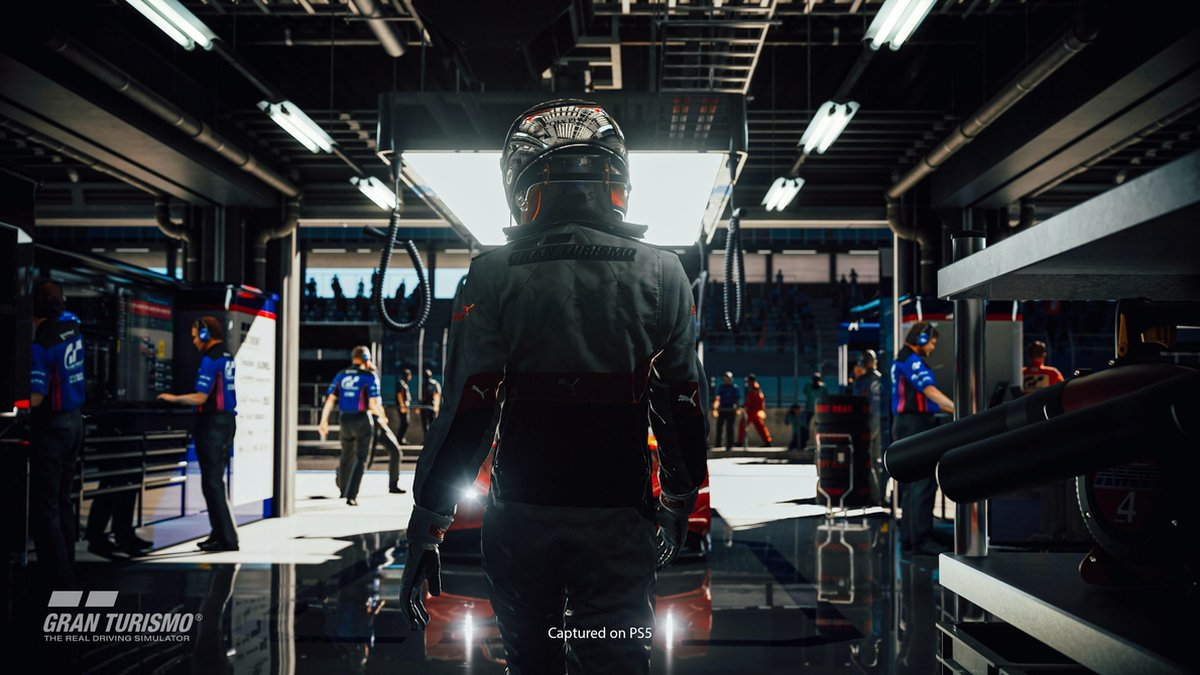 A person in racing gear stands in a garage, looking towards a race track. Other people are present.