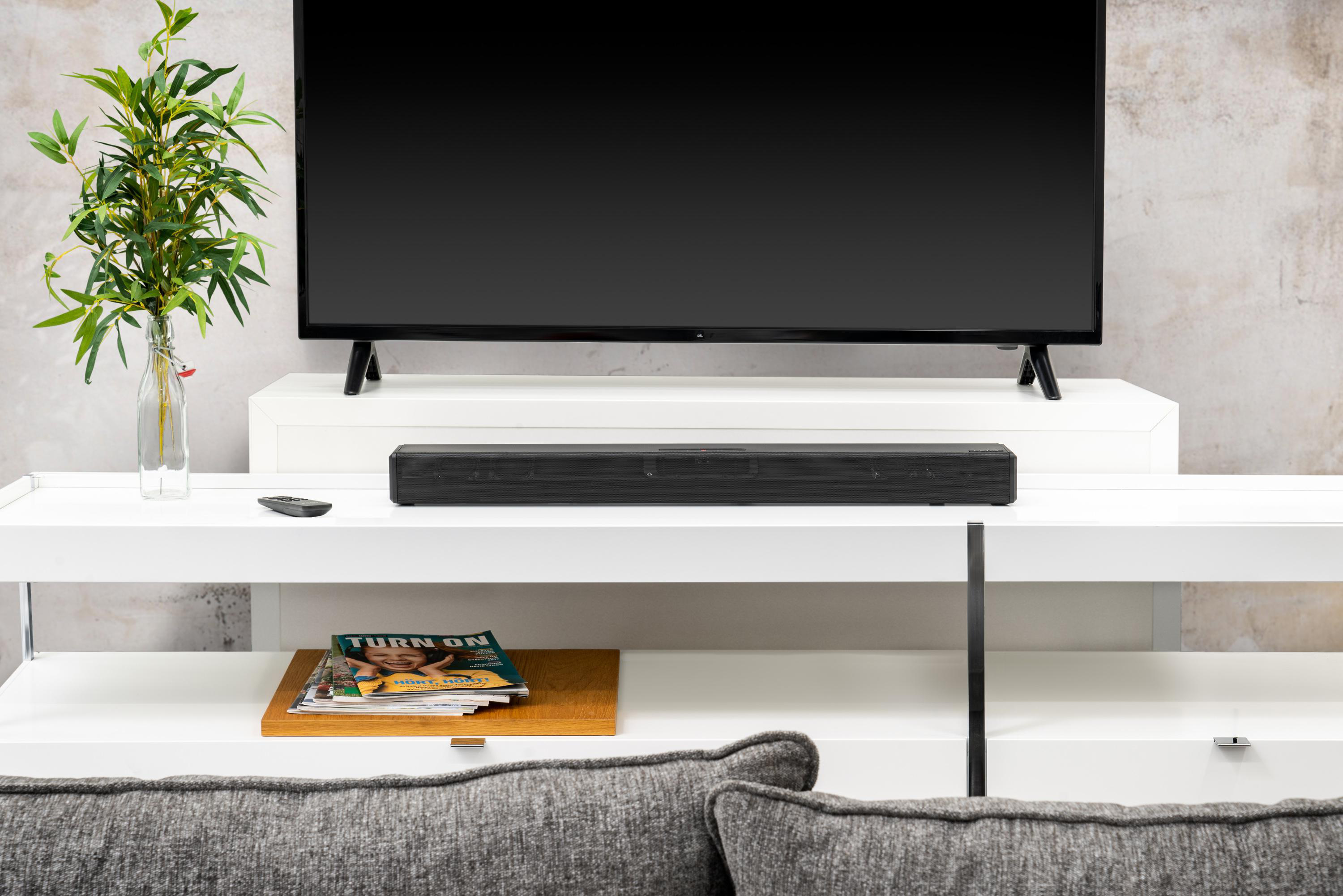 Wohnzimmer mit Fernseher auf einem weißen Regal, Soundbar darunter und Zeitschriften. Grüne Pflanze links.
