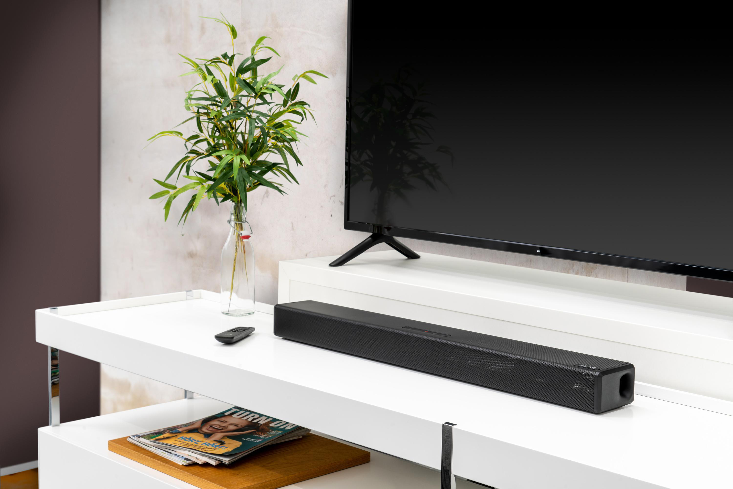 Schwarze Soundbar auf weißem Regal. Im Hintergrund ein Fernseher. Pflanzen und Fernbedienung.