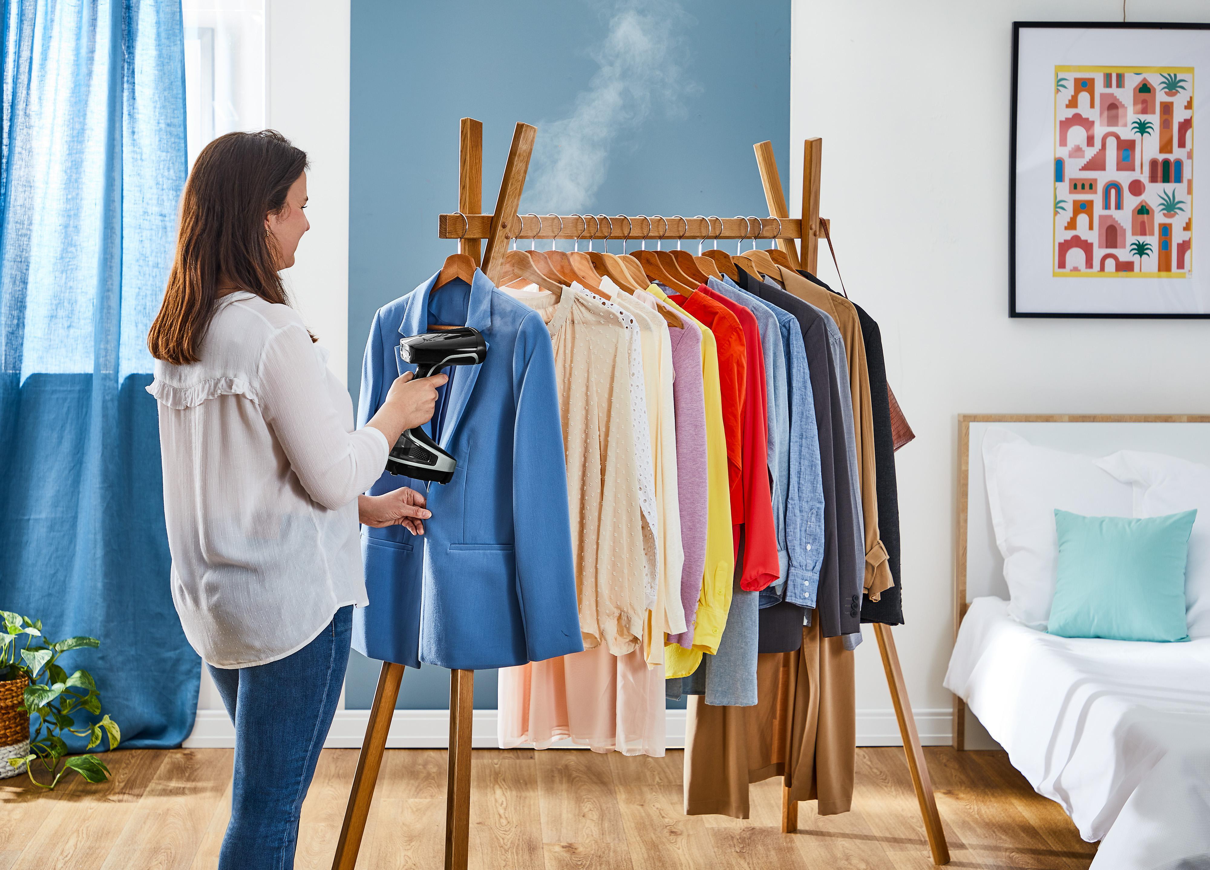 Frau, die eine blaue Jacke auf einem Holzgestell bügelt, mit Kleidung und Bett.