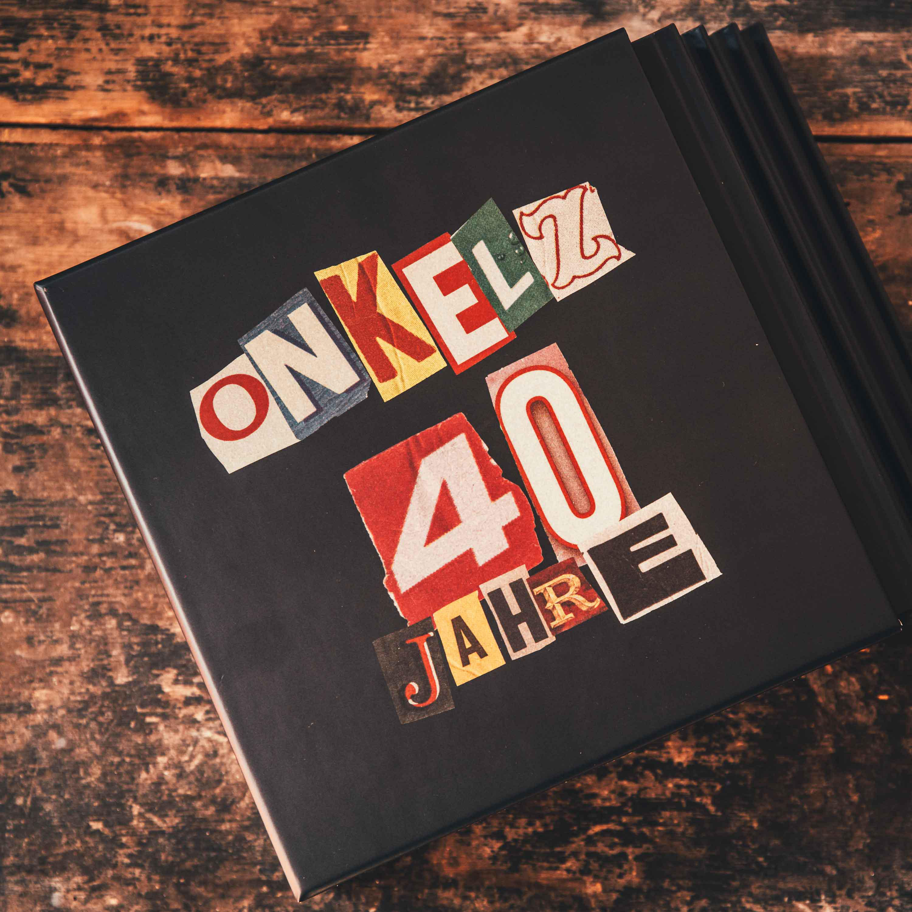 Ein schwarzes Buch mit einer bunten Collage, die "ONKELZ 40 JAHRE" sagt, auf einer Holzoberfläche.