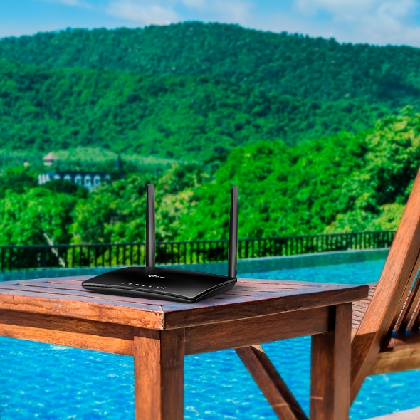 A black router sits on a wooden table with two antennas. The background is a pool and mountains.