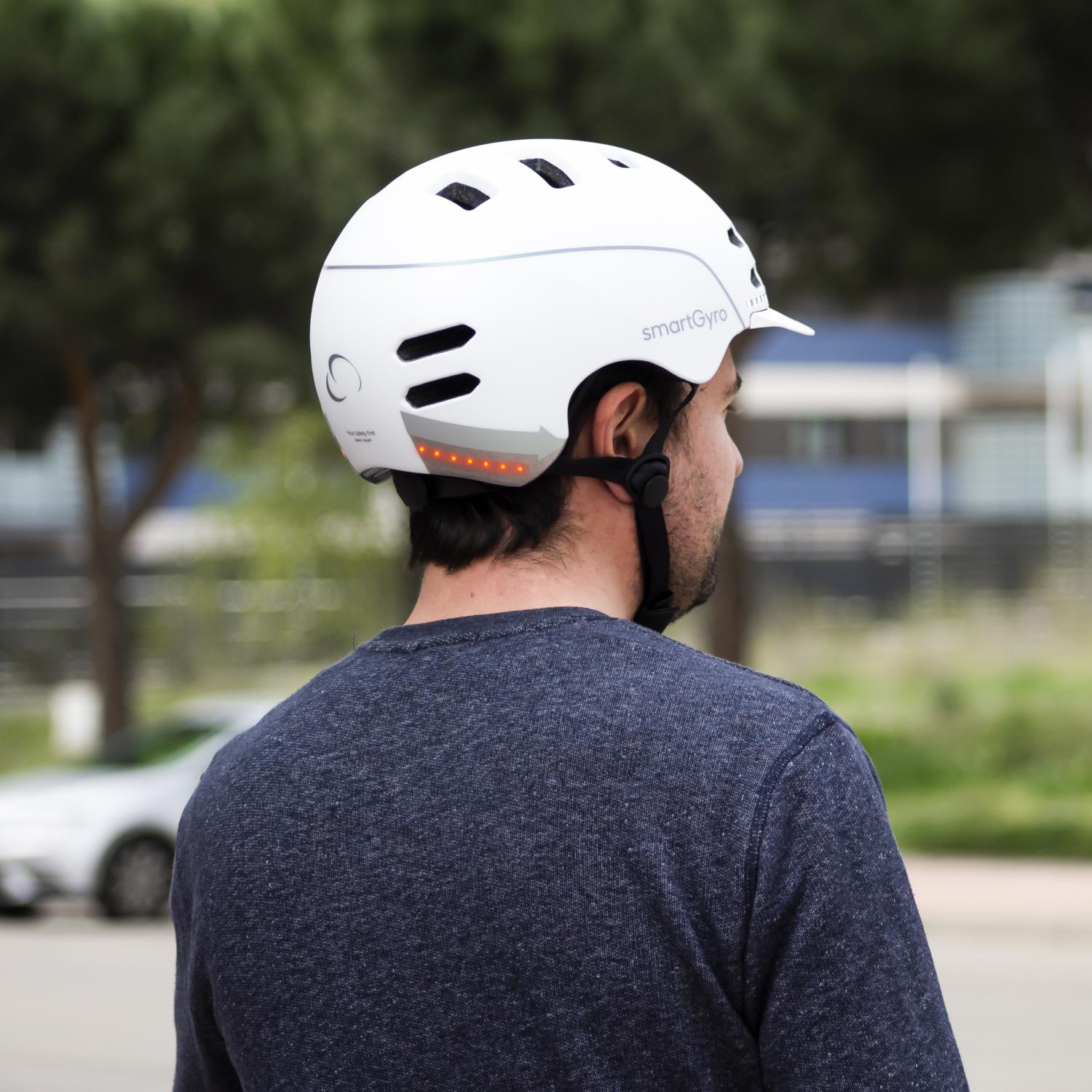 Un hombre con un casco blanco y una correa negra. Está de pie al aire libre con árboles y un coche al fondo.