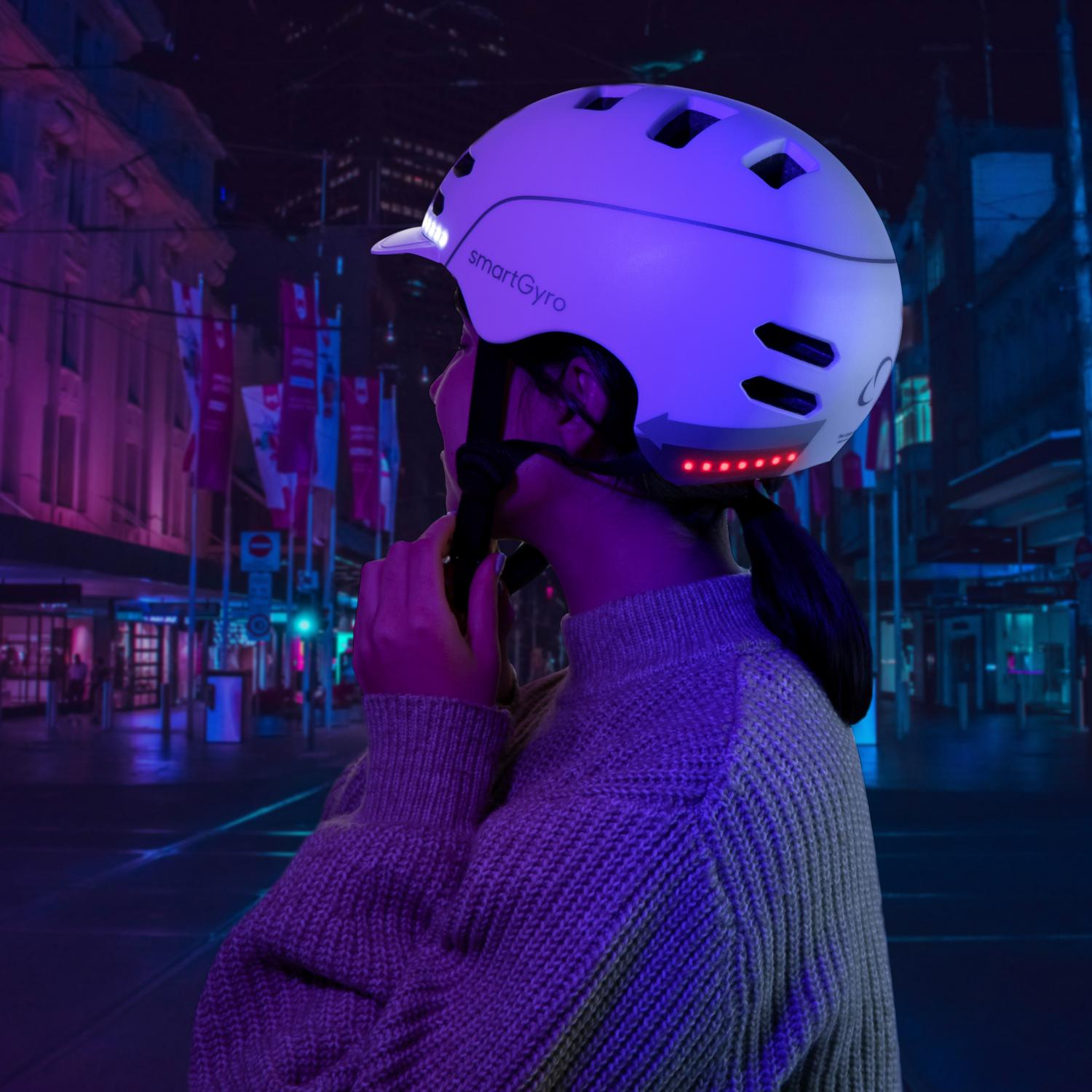 Persona con casco smartGyro blanco por la noche. Fondo urbano, luces.