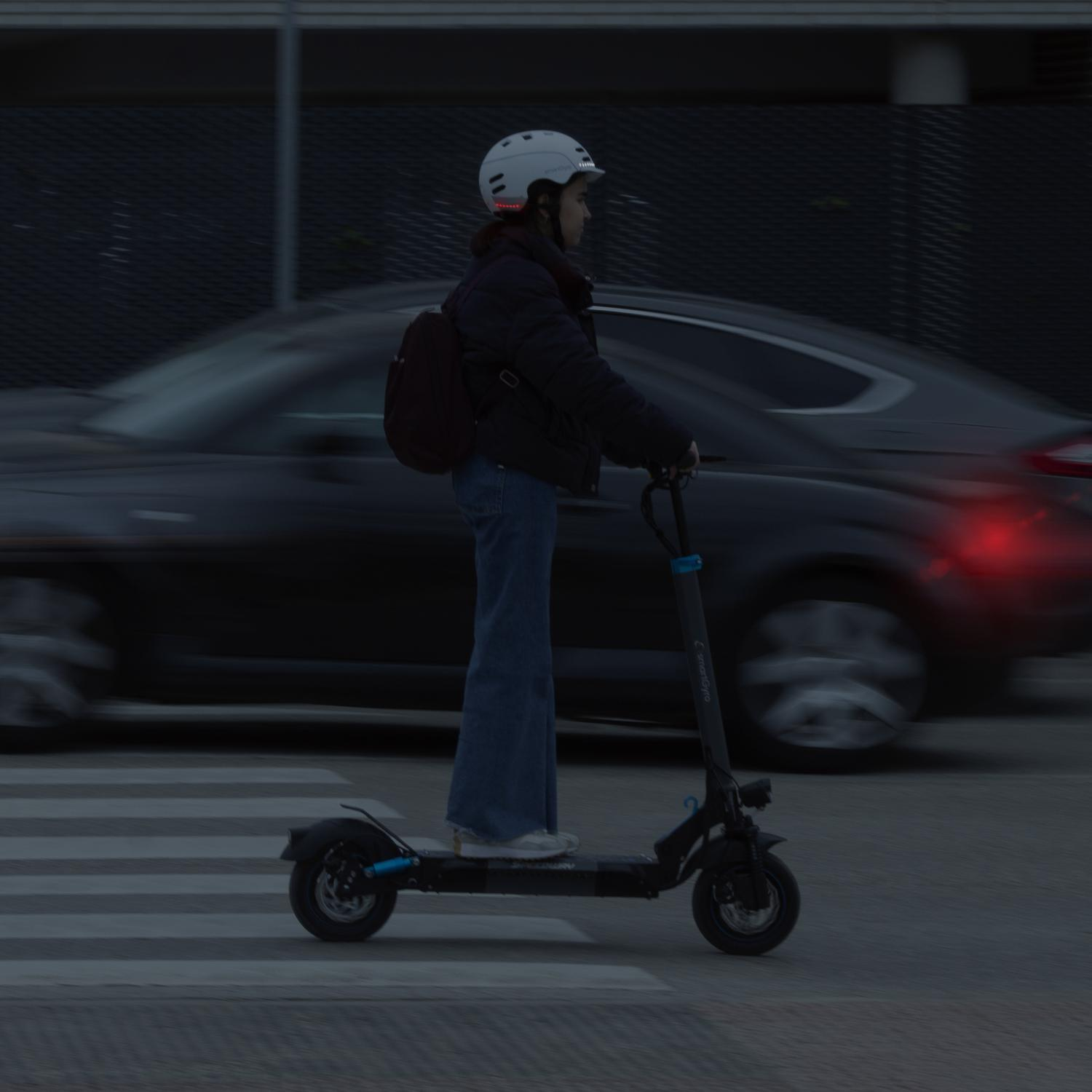 Persona monta un scooter en un paso de cebra por la noche, coche al fondo.