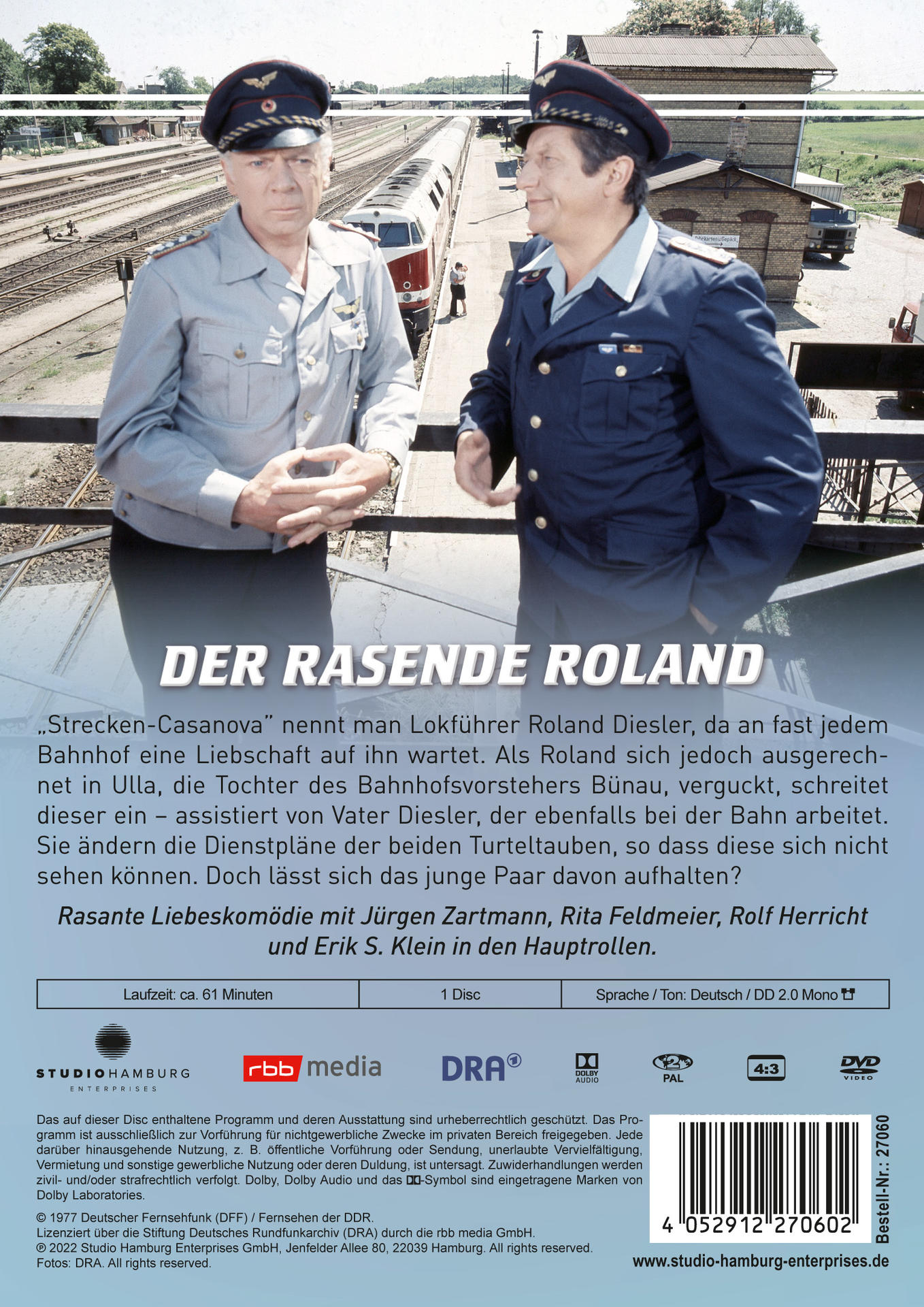 Zwei Männer in Uniform stehen auf einem Bahnsteig mit einem Zug und einem Gebäude im Hintergrund.