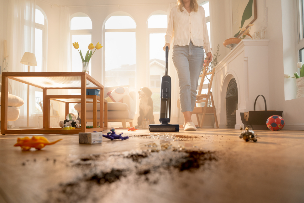 Eine Frau saugt ein unordentliches Wohnzimmer. Spielzeug und Schmutz sind verstreut. Sonnenlicht scheint durch die Fenster.