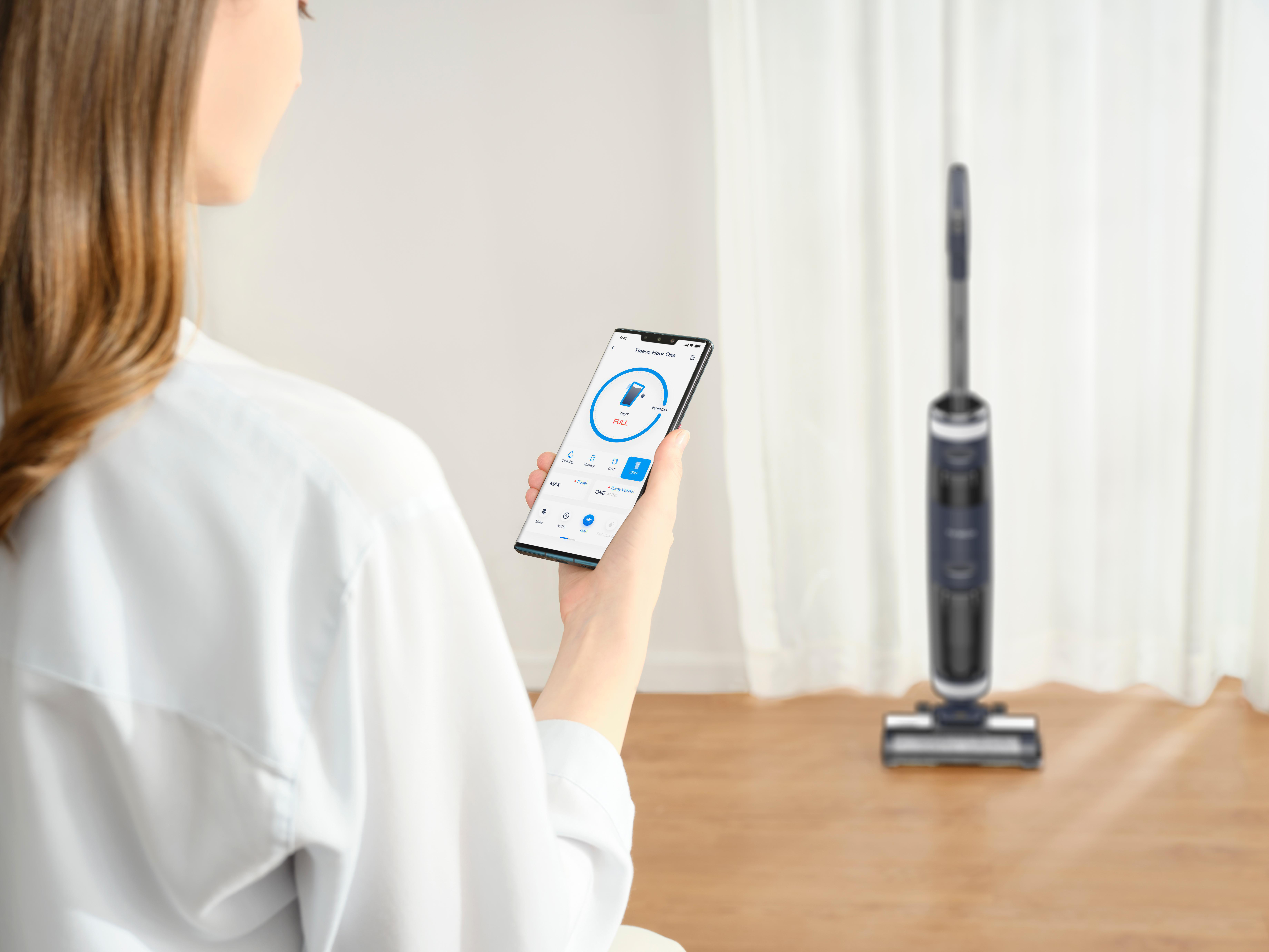 Frau hält Telefon mit Smart Home App, Staubsauger im Hintergrund.