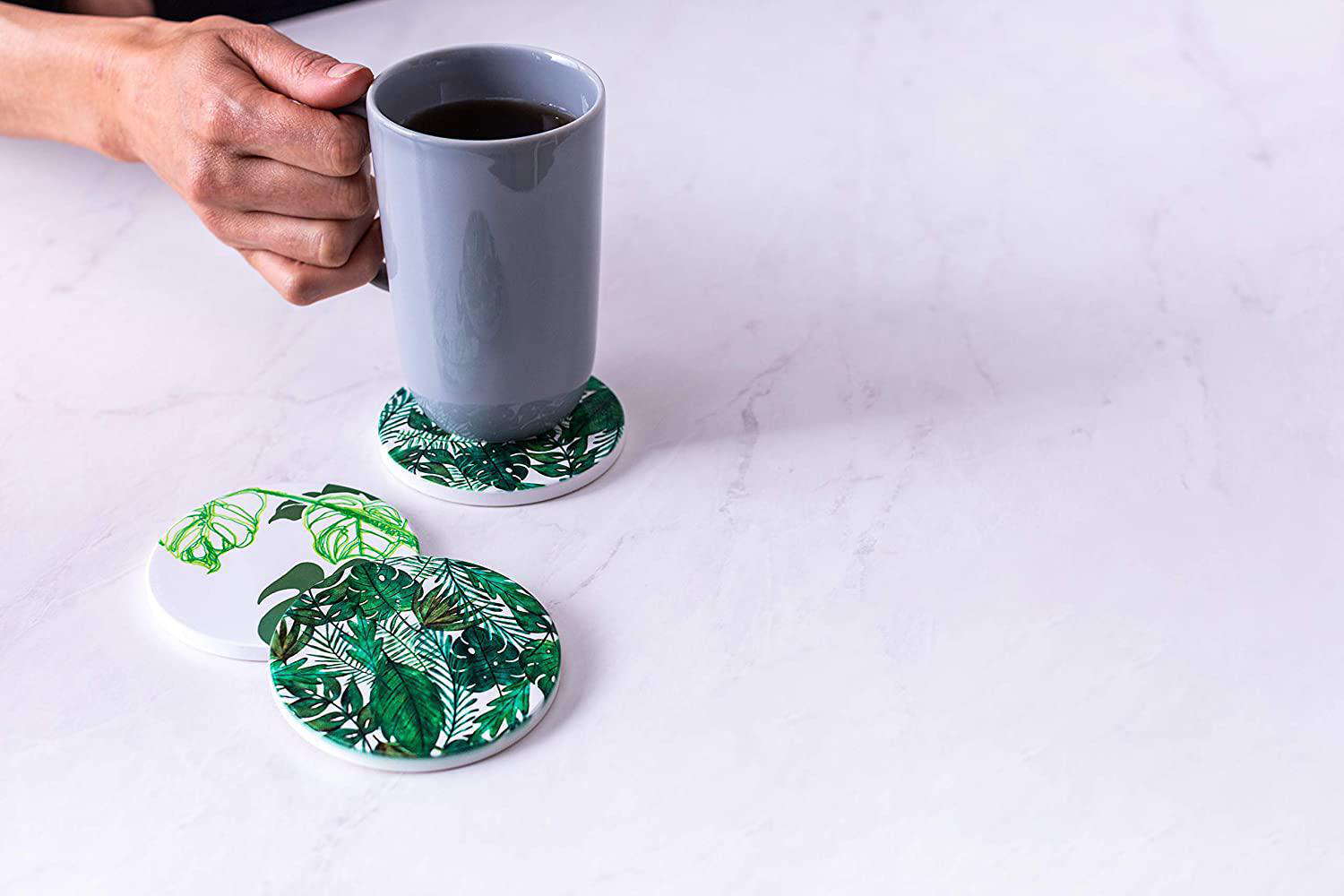 Hand, die einen grauen Becher auf einem weißen Tisch mit Blättermustern hält.