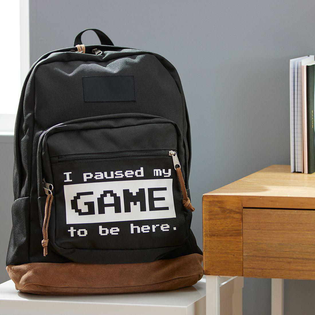 Schwarzer Rucksack mit dem Text "I paused my GAME to be here.", auf einem weißen Tisch, neben einem Holzschreibtisch.