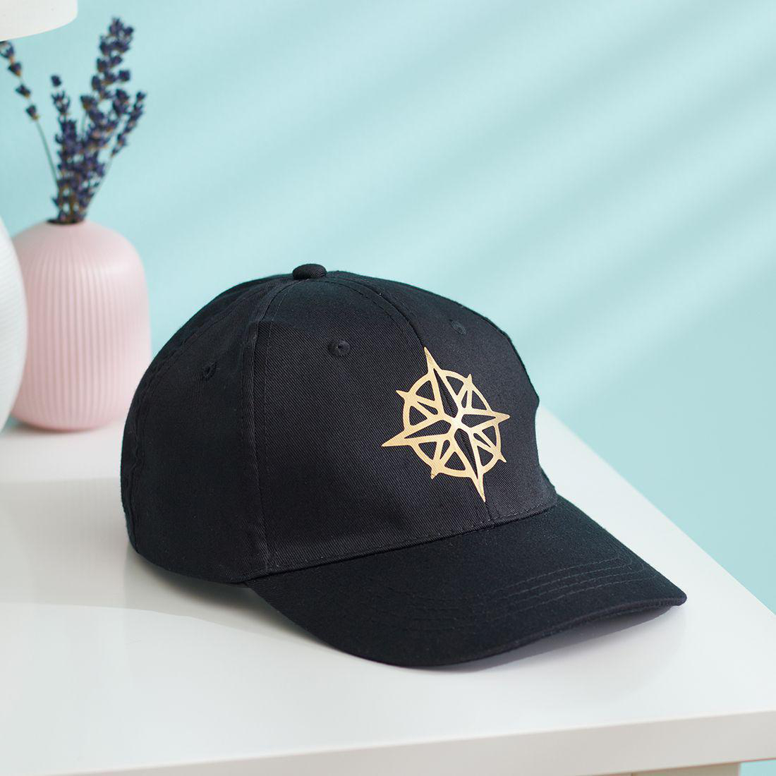 Eine schwarze Baseballkappe mit goldenem Kompassdesign liegt auf einem weißen Tisch. Eine rosa Vase und Lavendel sind im Hintergrund.