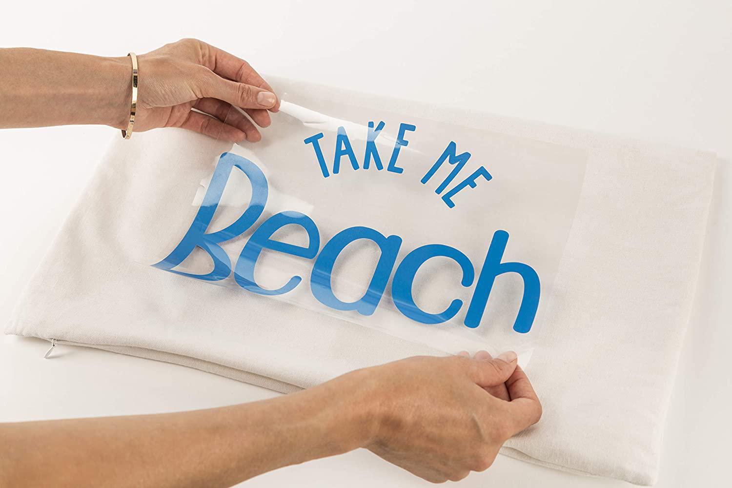 Hände, die einen blauen „Take Me Beach“-Aufkleber von einem weißen Kissen abziehen. Goldarmband sichtbar. Der Hintergrund ist schlicht weiß.