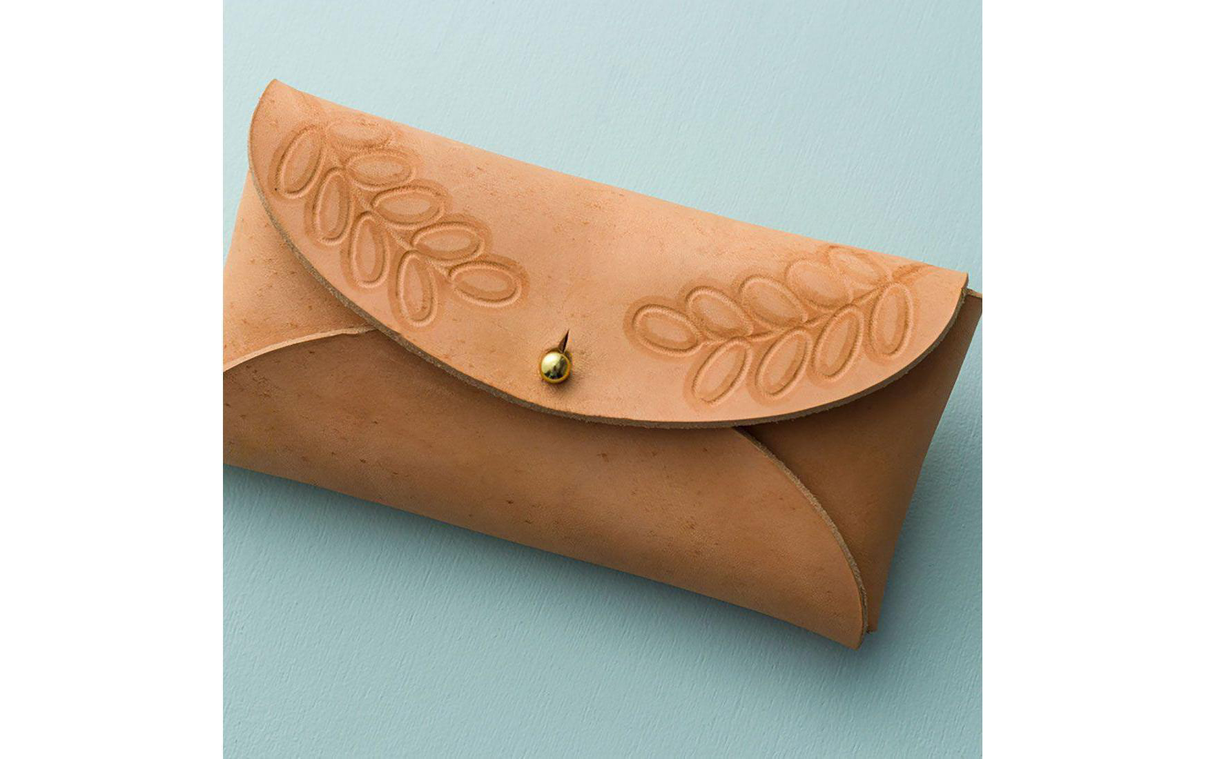 Eine hellbraune Lederclutch mit Blattmustern und einem goldenen Knopf. Sie liegt auf einer hellblauen Oberfläche.