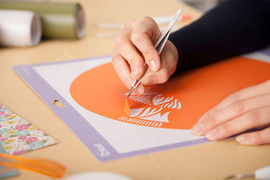 Die Hand einer Person verwendet eine Pinzette, um einen orangenen Aufkleber von einer Schneidematte zu ziehen.