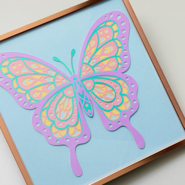 Ein gerahmter Papierfalter mit geschichteten Flügeln. Farben: Lila, Pink, Gelb und Türkis auf hellblauem Hintergrund.