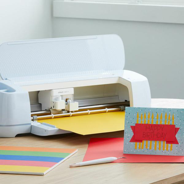 Eine Cricut-Maschine mit gelbem Papier, eine Geburtstagskarte und Bastelmaterialien stehen auf einem Holztisch.