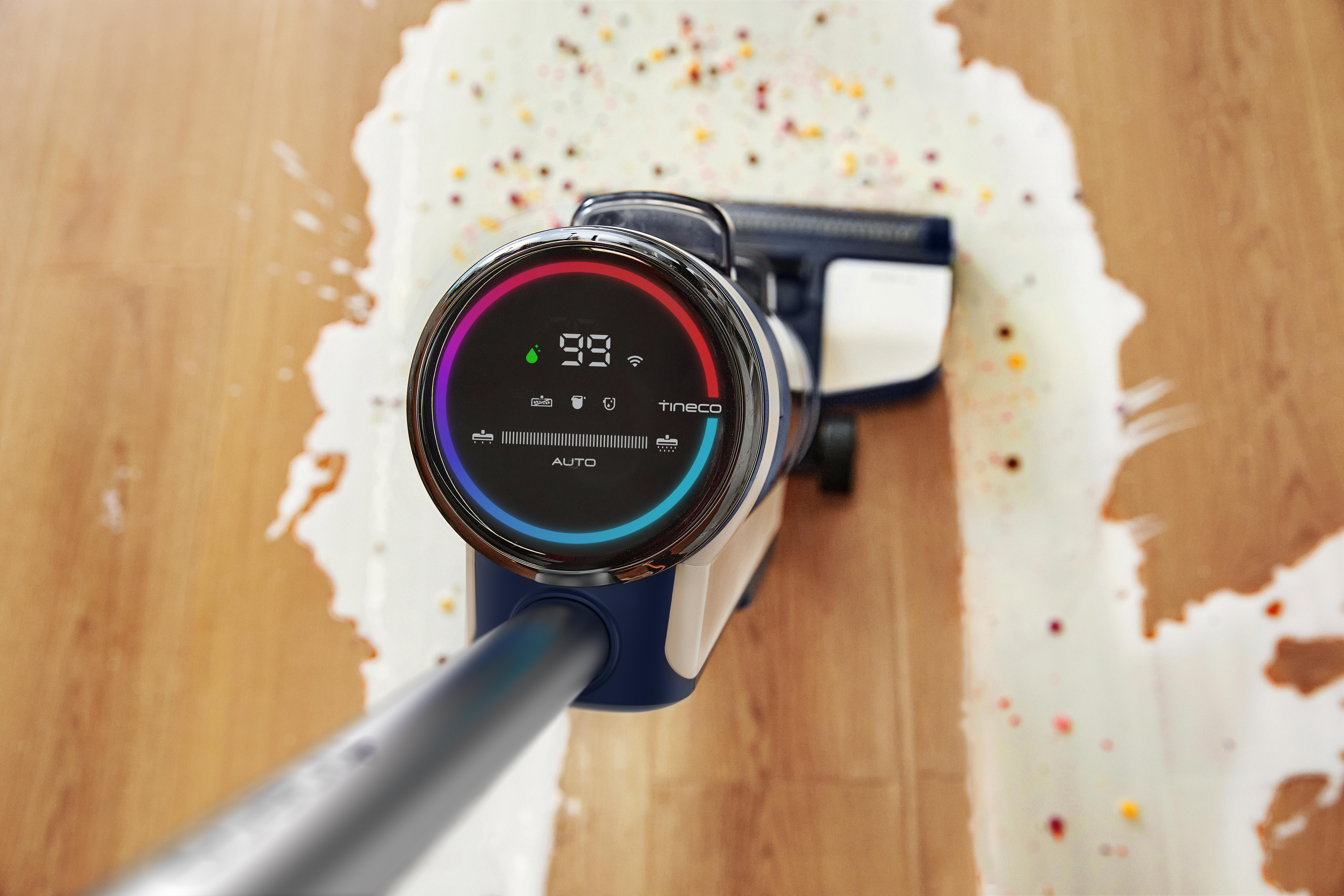 Ein Staubsauger mit Digitalanzeige, der einen Holzboden mit verschütteter Flüssigkeit reinigt.