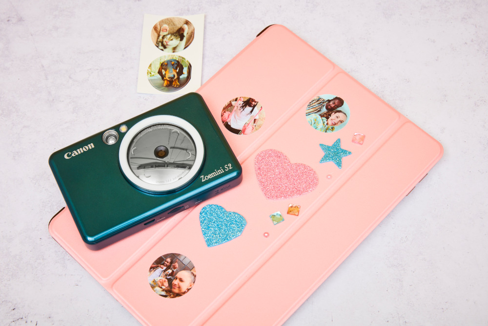 A green Canon Zoemini S2 camera, a pink tablet, and stickers. The items are on a white surface.