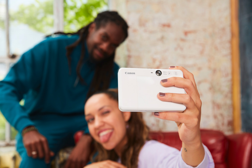 Two people taking a selfie with a white camera, one winking and sticking out tongue.