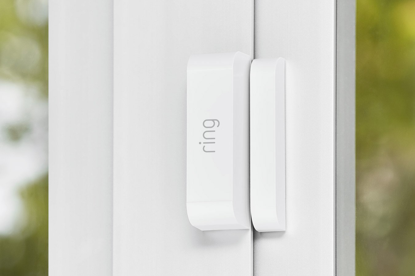 A white Ring door sensor is attached to a white door. The background is blurred green.