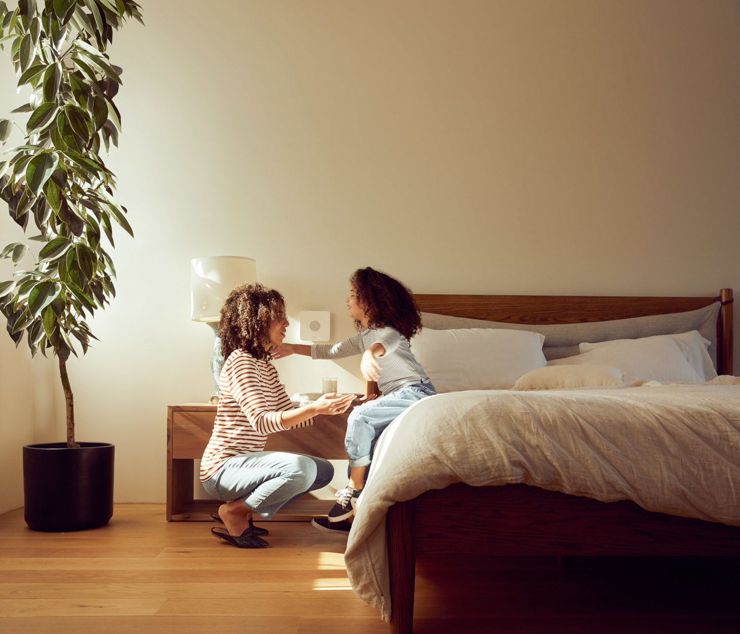 A woman and a child are near a bed. The woman is squatting, and the child is sitting on the bed. A plant is on the left.