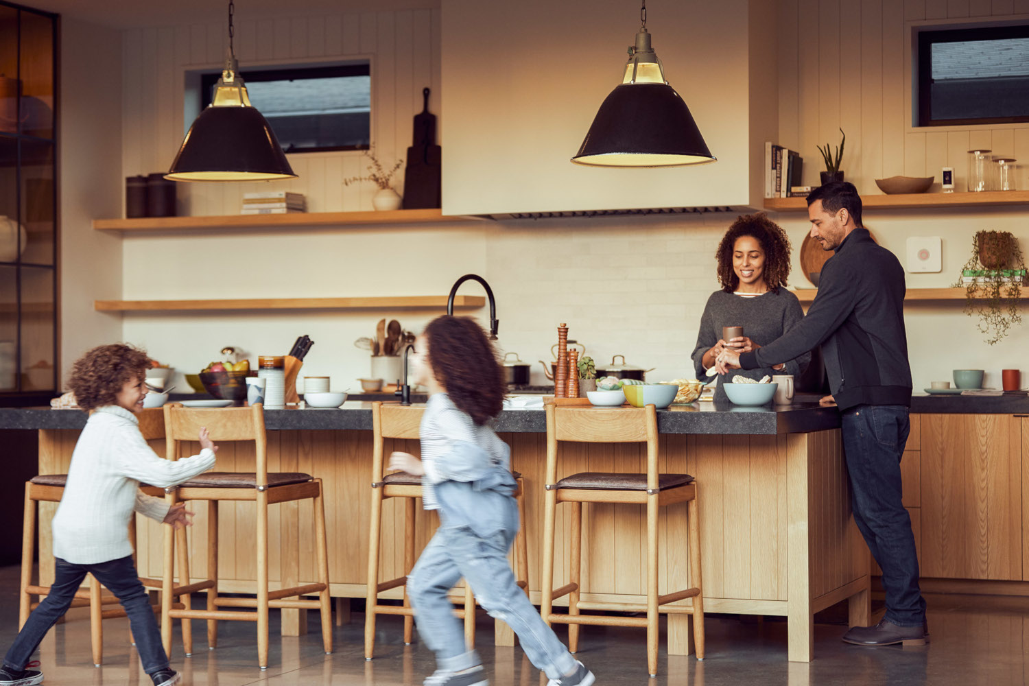 Family in a modern kitchen. Two kids run past the island counter. Parents stand behind the counter, preparing food.