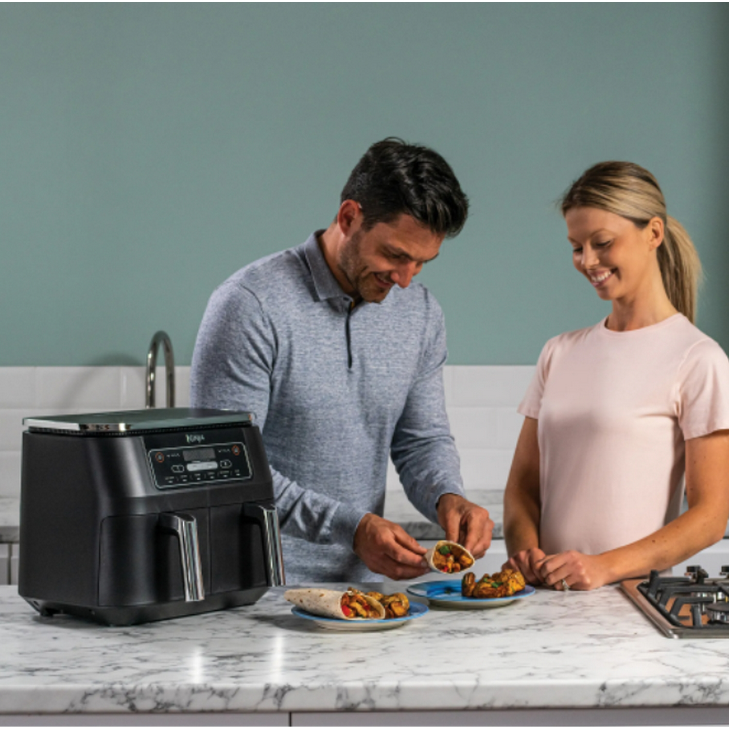 Pareja preparando comida con freidora de aire. Aparato negro, platos con comida. Ambiente de cocina.