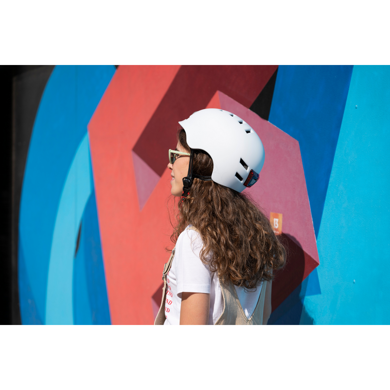 Mujer con casco blanco y gafas de sol de pie frente a una pared abstracta colorida.