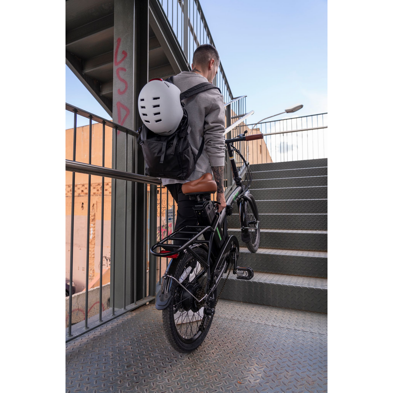 Una persona empujando una bicicleta negra por unas escaleras metálicas, llevando una mochila y un casco.